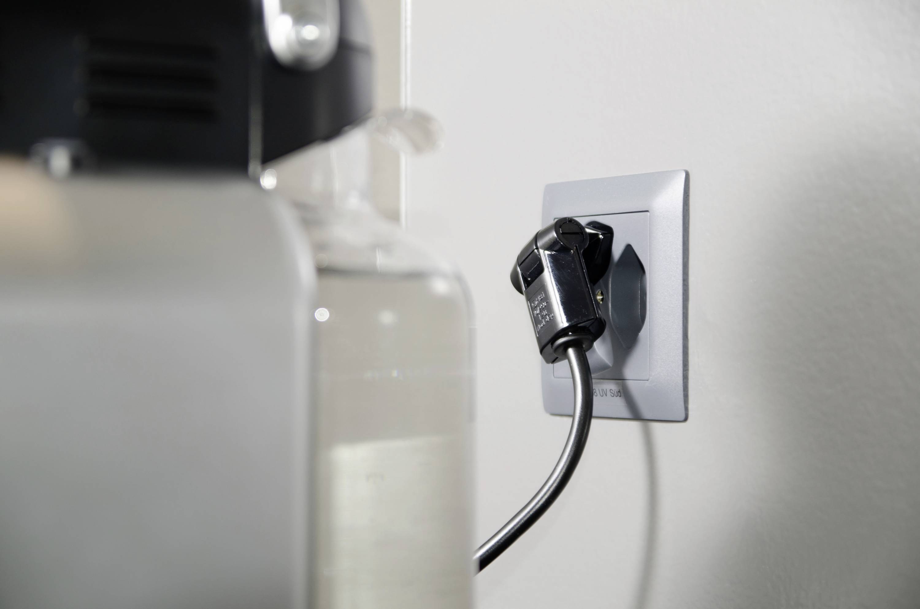 Ein elektrisches Gerät ist an eine Steckdose angeschlossen. Die Wand ist weiß, und das Kabel ist schwarz.