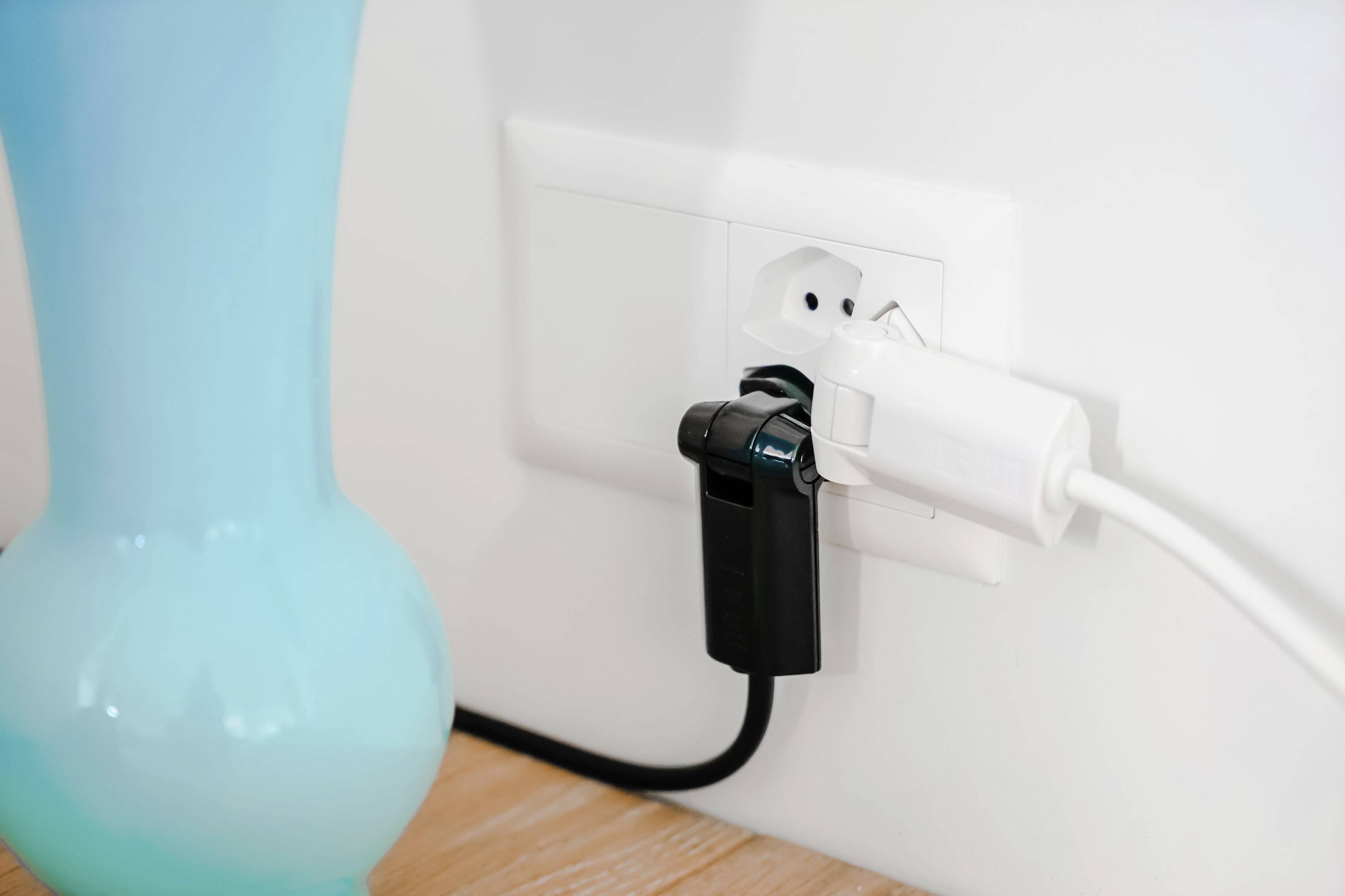 Ein schwarzes und weißes Ladegerät sind in eine weiße Steckdose eingesteckt. Links steht eine blaue Vase auf einem Holztisch.