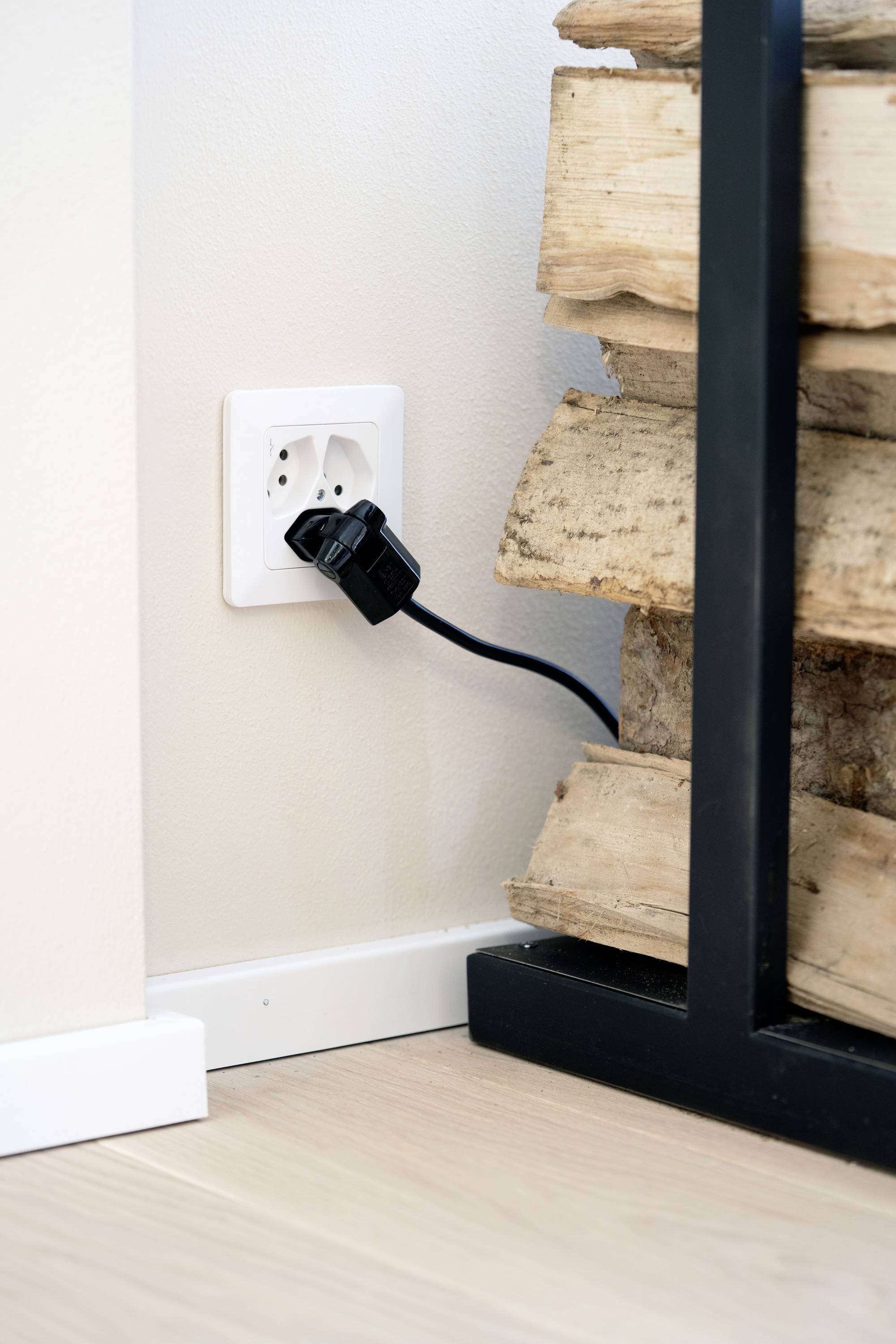 Steckdose in einer Wand mit eingestecktem Stecker, daneben gestapelte Holzscheite, Innenaufnahme in einer Ecke eines Raumes.