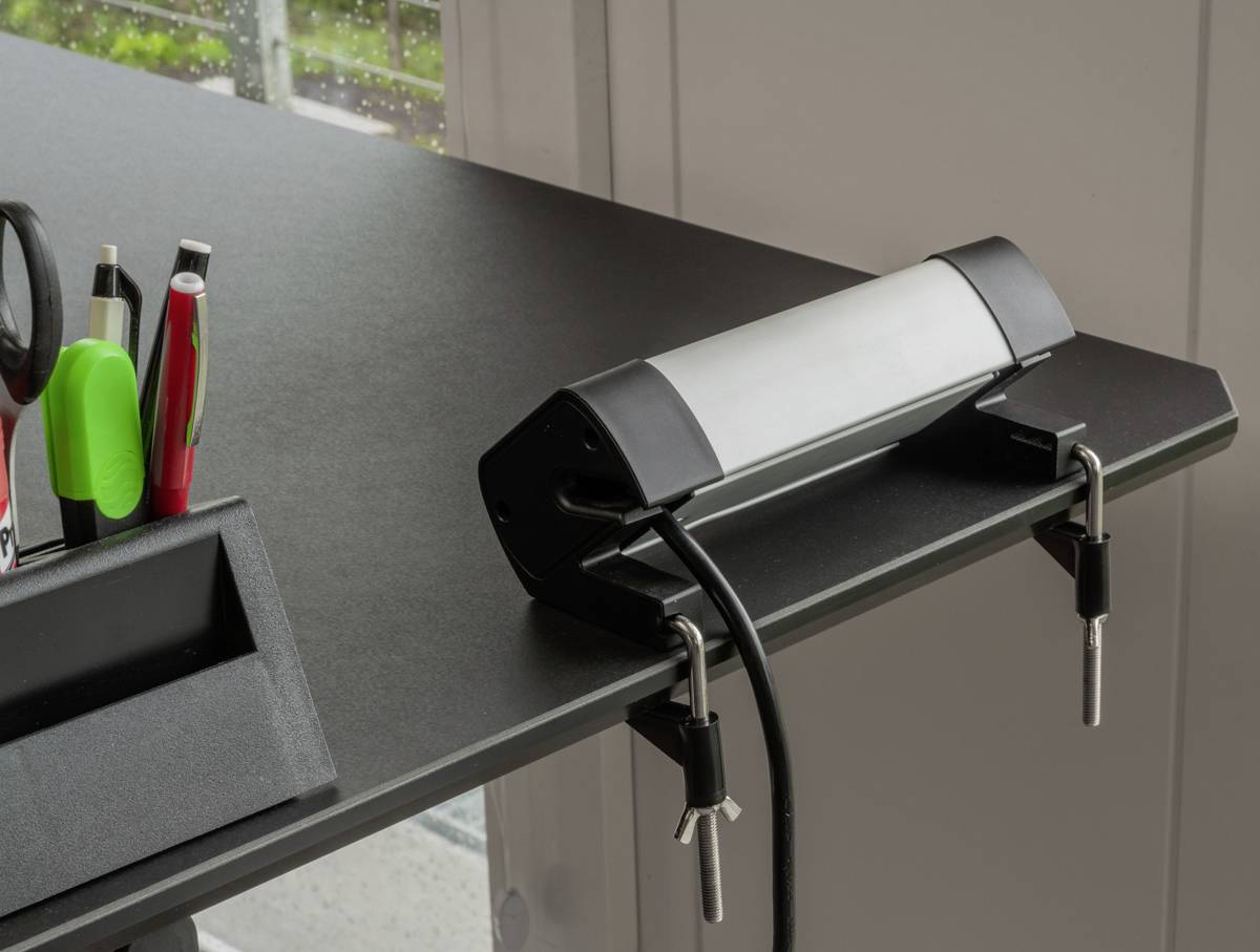 Eine schwarze Schreibtischlampe mit Clips ist an einer Tischkante befestigt, daneben stehen Stifte in einem Halter.