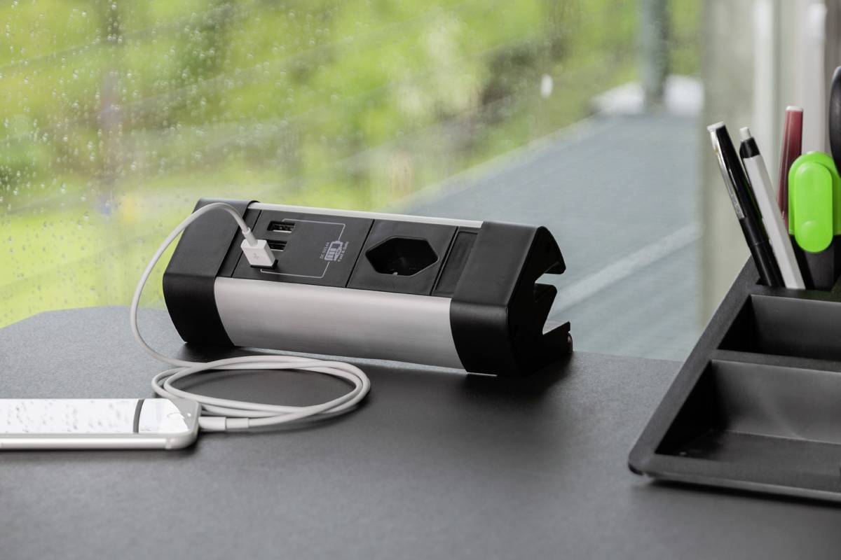 Ein Smartphone wird über ein USB-Kabel an eine Tischsteckdosenleiste angeschlossen. Im Hintergrund ist ein Fenster mit Blick nach draußen.