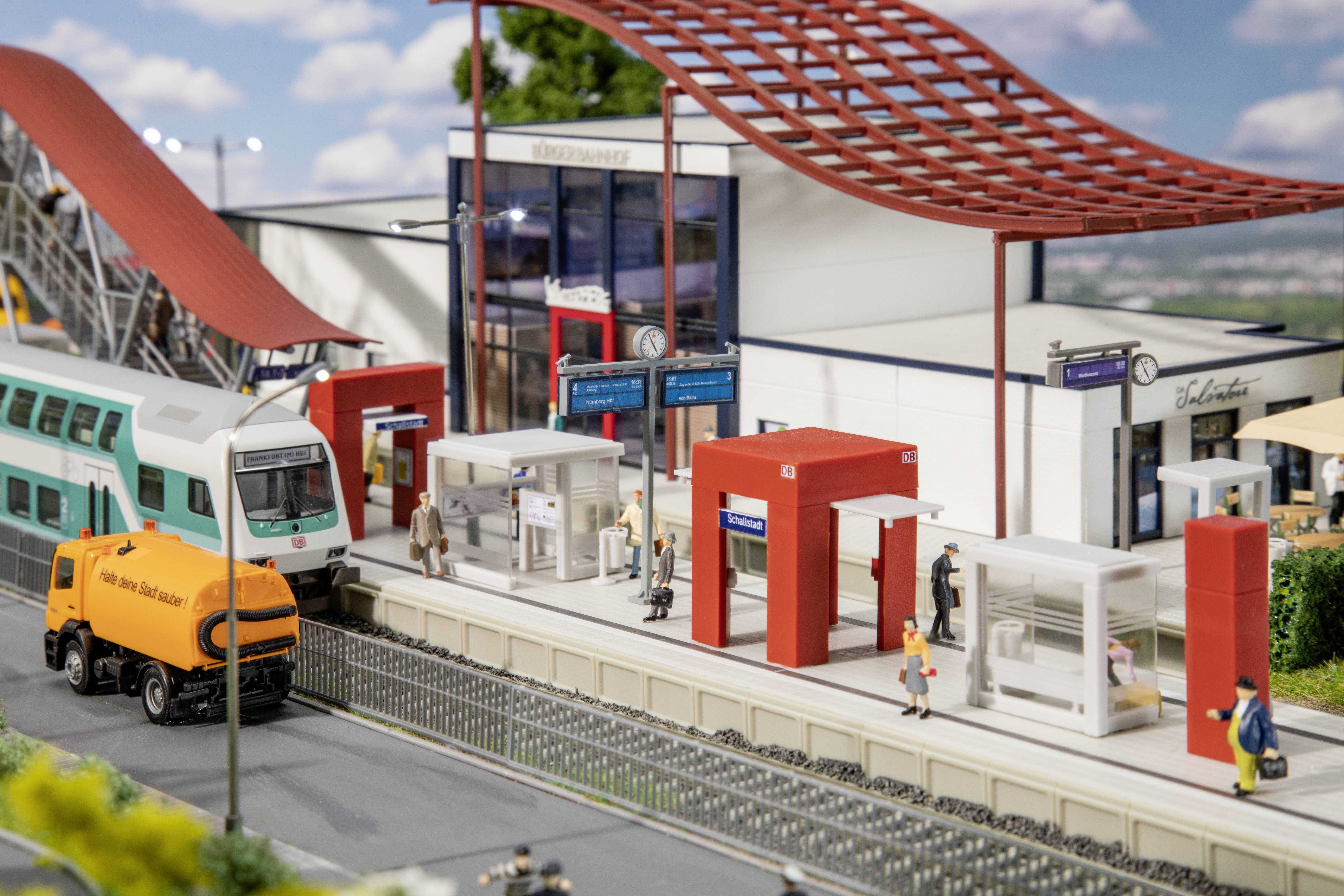 Ein Modellbahnhof mit einem Zug auf Gleis, umgeben von Figuren und einem orangefarbenen Fahrzeug. Moderne Architektur mit rotem Dach.