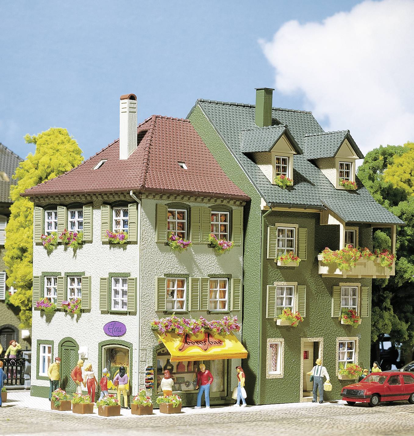 Mehrstöckiges Gebäude mit Schaufenster und Markise im Erdgeschoss. Menschen spazieren vorbei. Pflanzkästen mit Blumen an Fenstern.