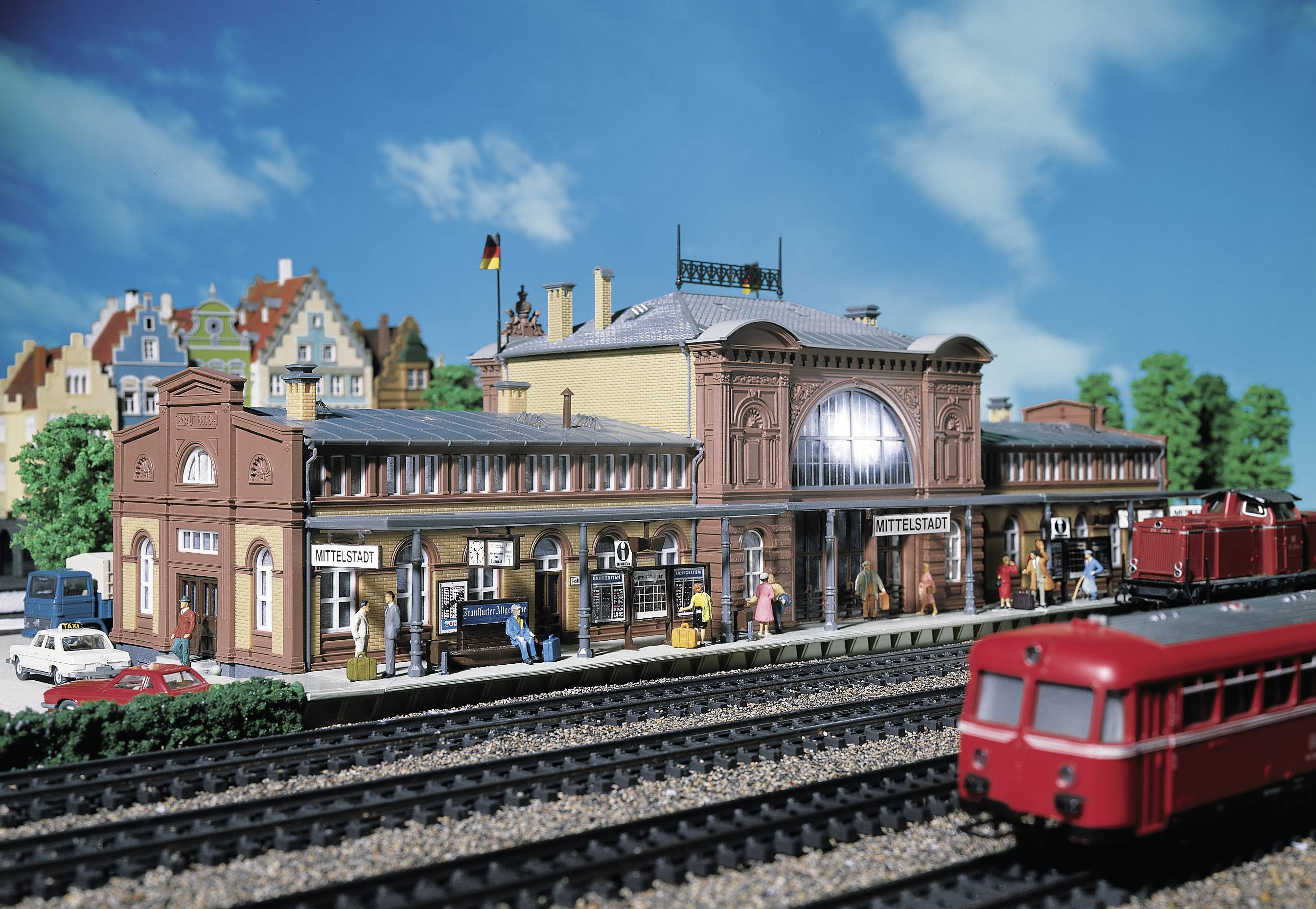 Modell eines Bahnhofs mit Zügen, Gleisen und Menschen in einer Miniaturstadt. Der Bahnhof ist mit Tischen und Schildern detailliert gestaltet.