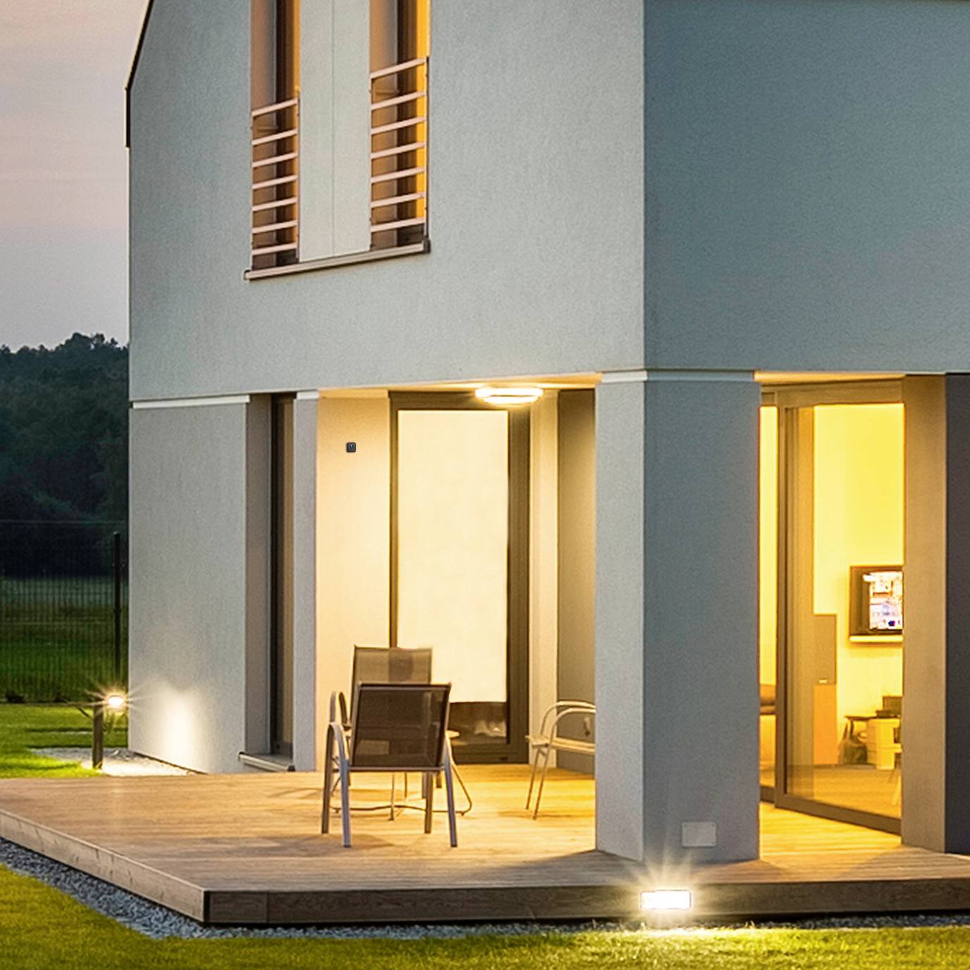 Modernes zweistöckiges Haus bei Dämmerung. Außenbeleuchtung erhellt den Eingangsbereich und die Terrasse, auf der Stühle stehen.