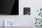 Flachbildfernseher an der Wand neben dekorativem Wandregal mit zwei Bilderrahmen, einer leer, einer mit Kunst. Pflanze rechts im Bild.