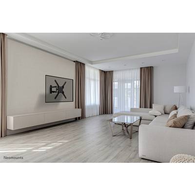 Wohnzimmer mit hellen Wänden und Holzboden. Ein großes Sofa mit Kissen steht vor einem TV-Wandhalter ohne Fernseher. Große Fenster mit langen Vorhängen lassen Licht herein.