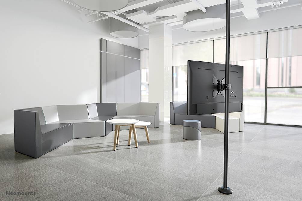 Ein moderner, heller Raum mit grauem Sitzbereich, zwei runden weißen Tischen und einem großen Bildschirm auf einem Standfuß.