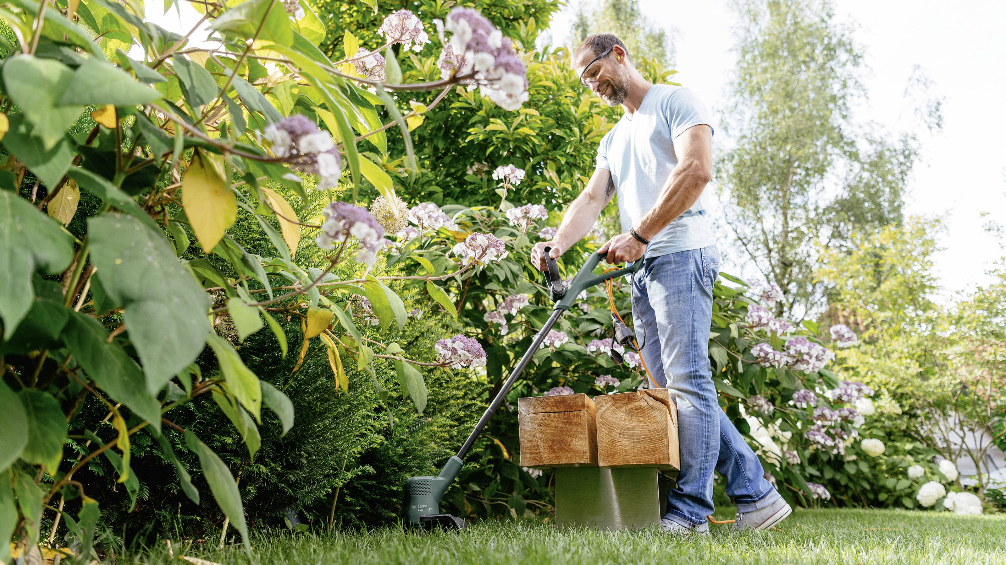 Ein Mann stutzt Gras in einem Garten mit einem Trimmer. Er trägt ein hellblaues Hemd und Jeans. Üppiges Grün und blühende Blumen umgeben ihn.