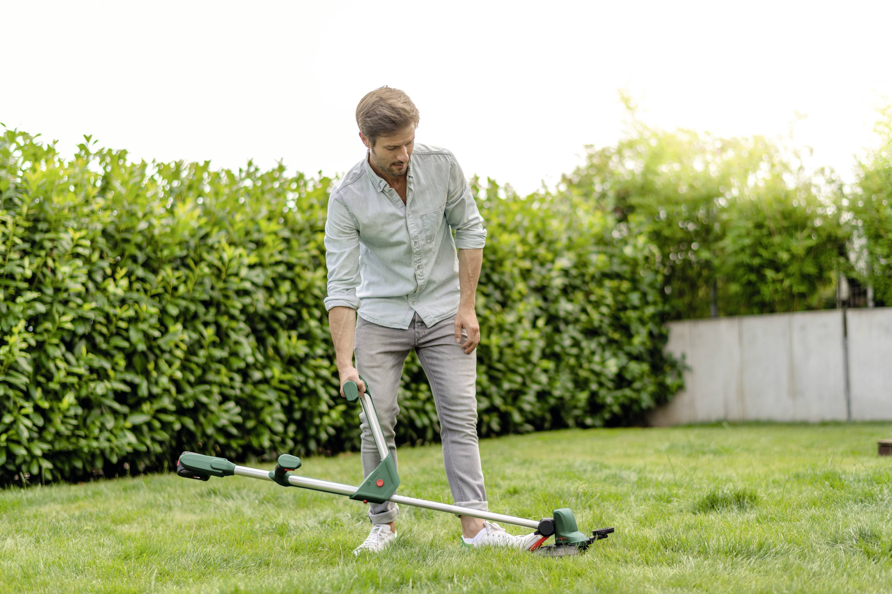 Ein Mann im Garten trimmt mit einem elektrischen Rasentrimmer das Gras. Im Hintergrund sind Hecken und ein Zaun zu sehen.