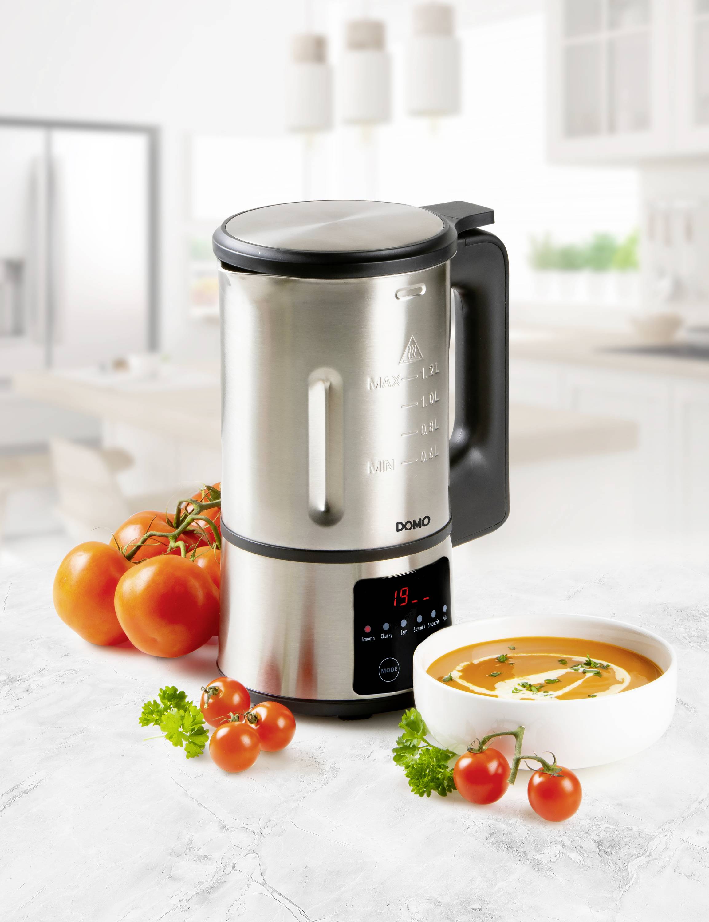 Ein moderner Suppenbereiter aus Edelstahl steht auf einer Küchenarbeitsplatte. Daneben liegen Tomaten, und eine Schüssel Suppe ist serviert.