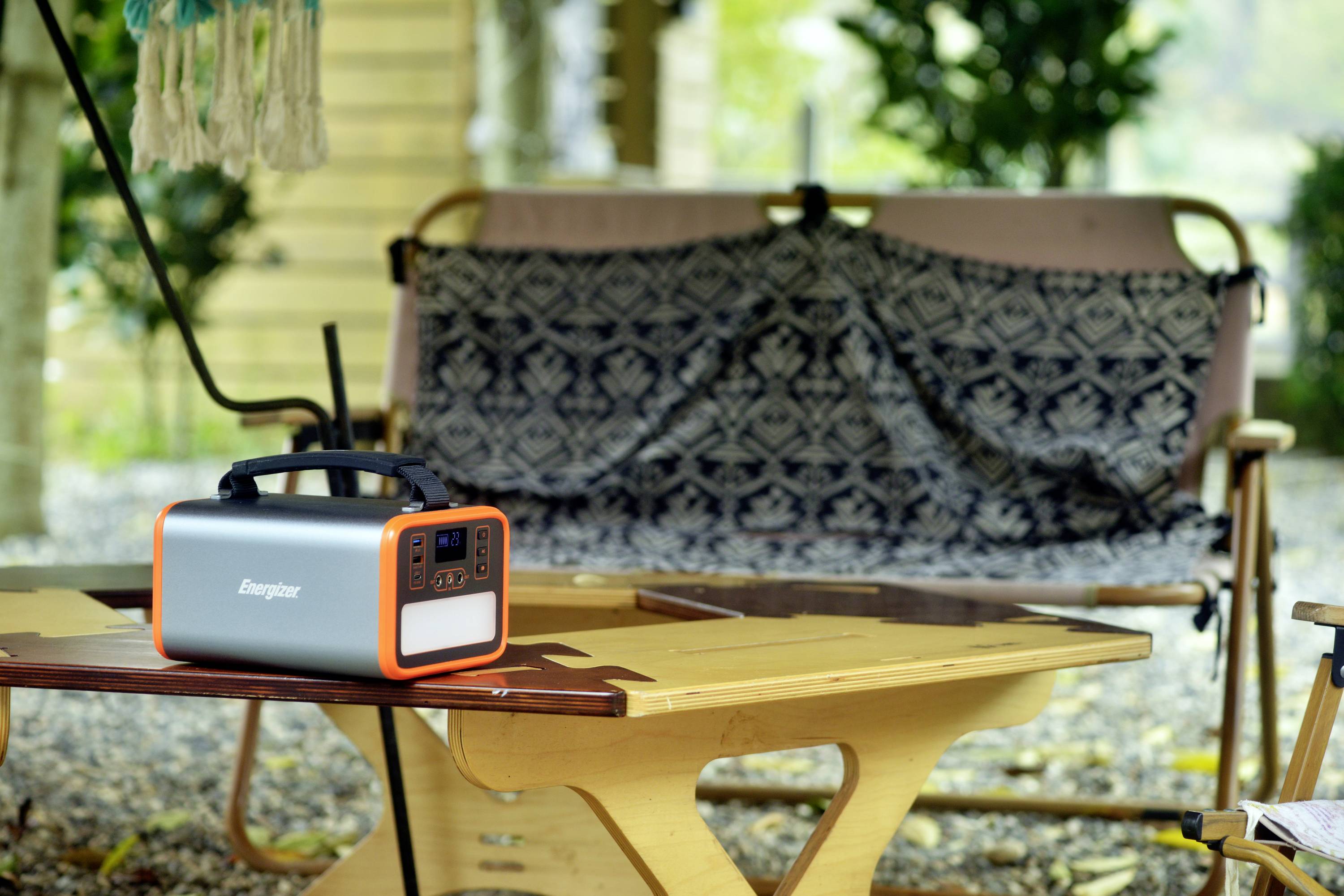 Tragbare Powerstation auf einem Holztisch im Freien, dahinter eine gefaltete Decke auf einem Stuhl. Umgebung wirkt entspannend und naturverbunden.