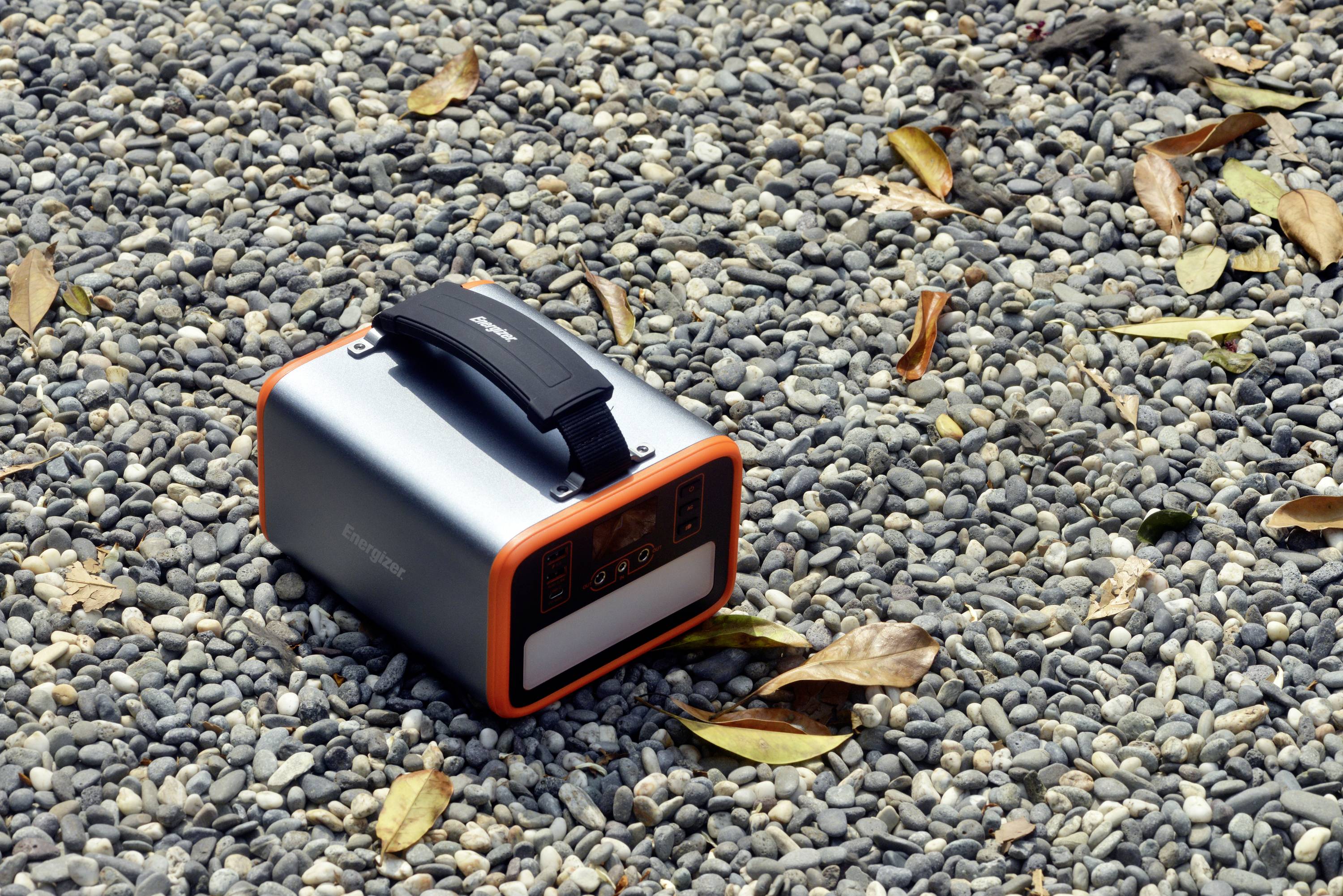 Kompakte tragbare Powerbank mit Tragegriff, auf einer Kiesfläche. Umgeben von herabgefallenen Blättern. Ideal für Outdoor-Aktivitäten.