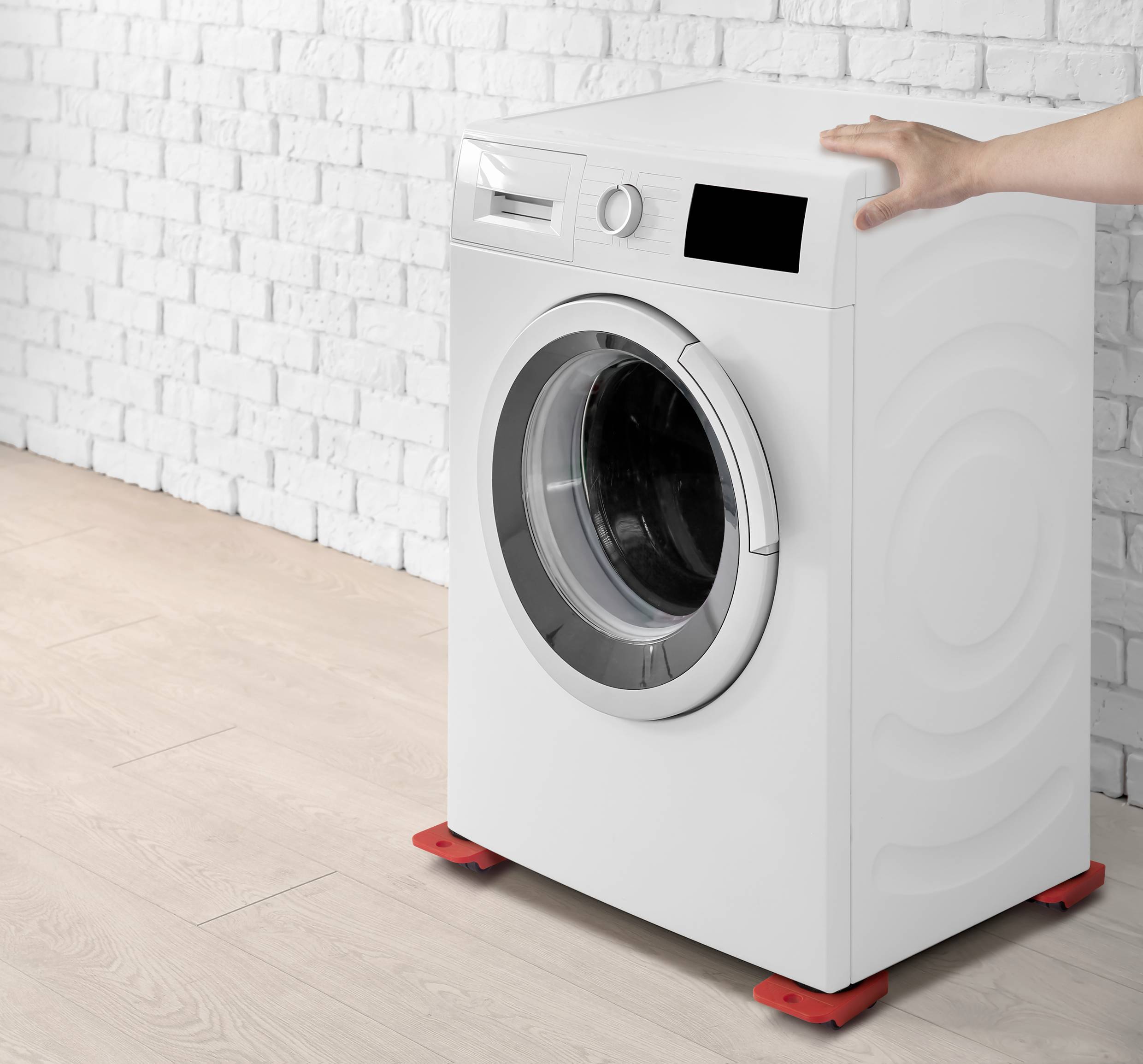Eine weiße Waschmaschine steht auf einem Holzboden vor einer weißen Backsteinwand. Eine Hand berührt die Oberseite der Maschine.