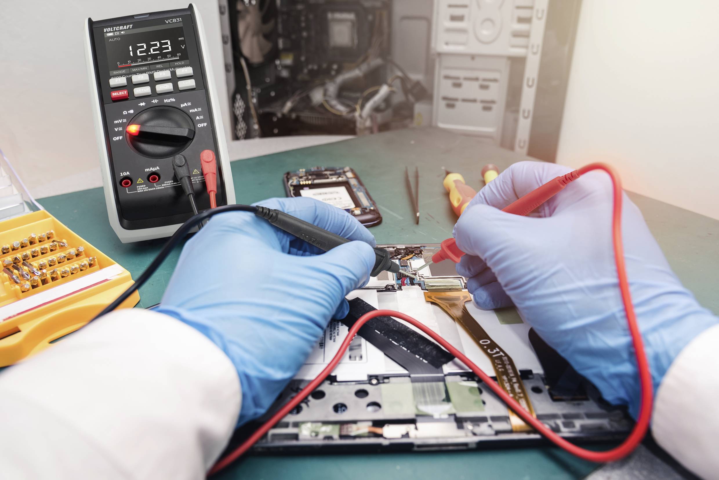 Eine Person repariert ein elektronisches Gerät mit einem Multimeter. Schraubenzieher und Platine sind auf dem Tisch zu sehen.