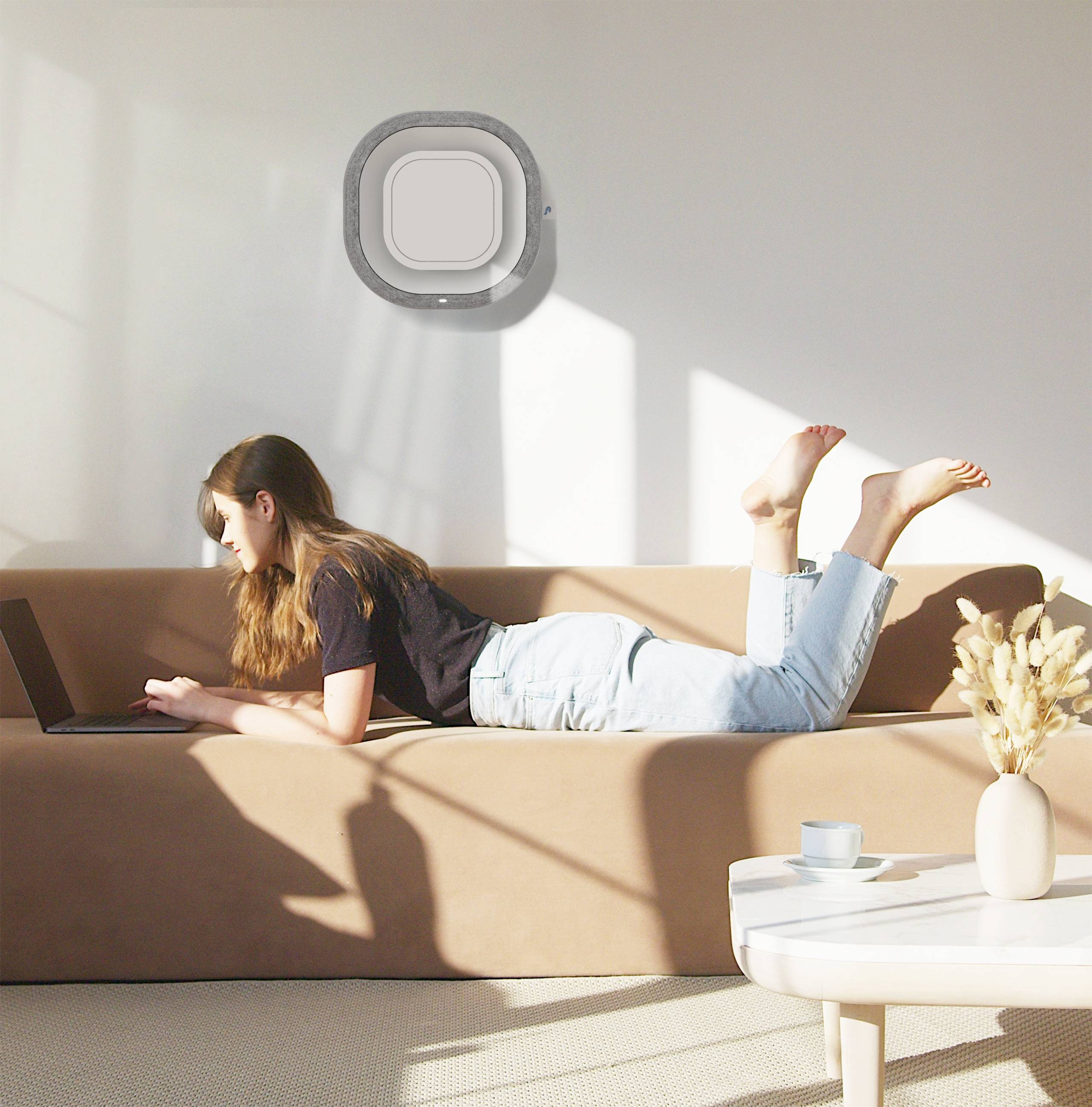 Eine Person liegt auf einem Sofa und arbeitet an einem Laptop. Sonnenlicht fällt durch ein Fenster. Ein Tisch mit einer Tasse steht im Vordergrund.