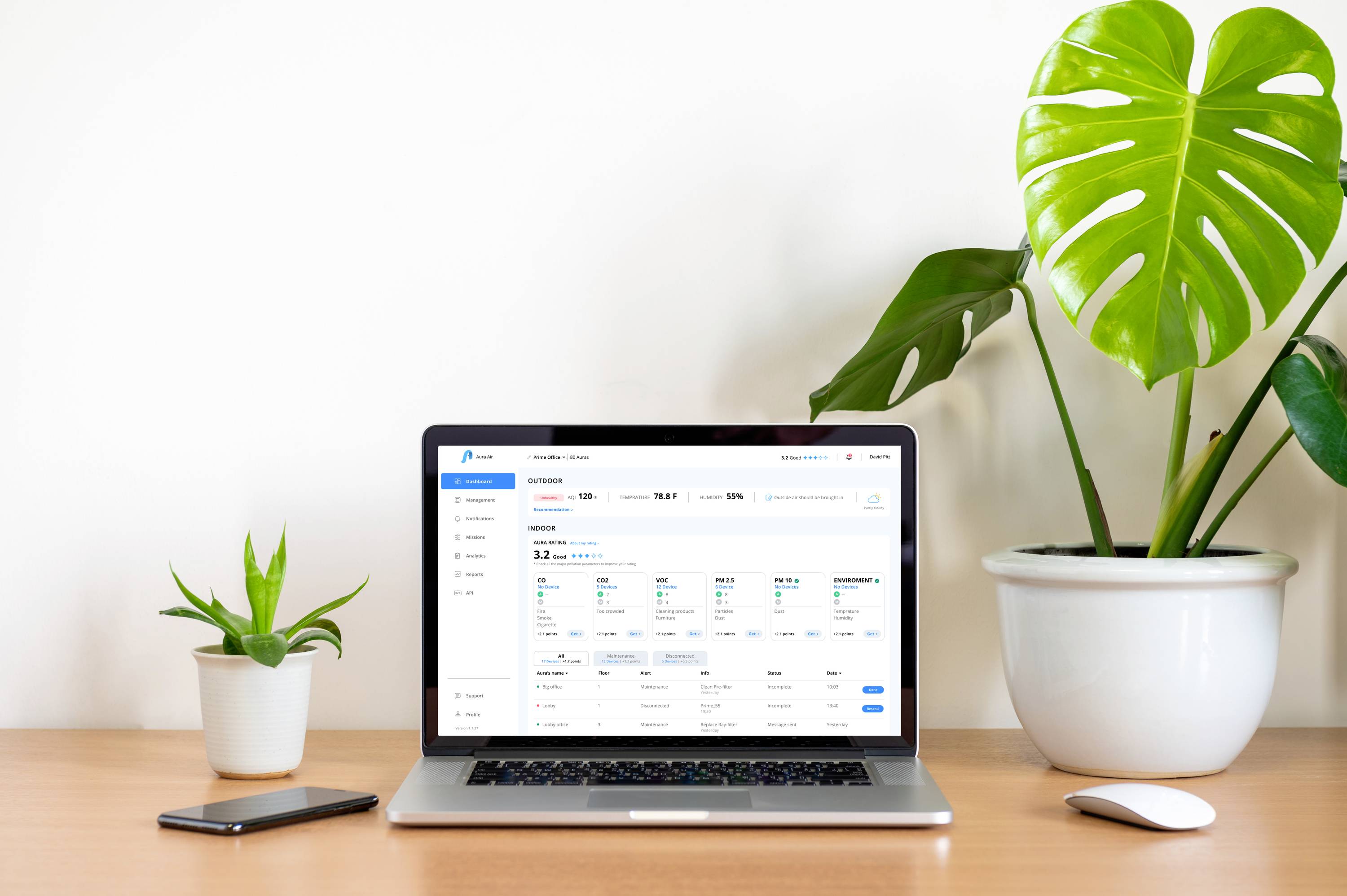 Ein Laptop zeigt eine geöffnete Website mit Finanzdaten auf einem Tisch zwischen zwei Pflanzen. Daneben liegt ein Smartphone.