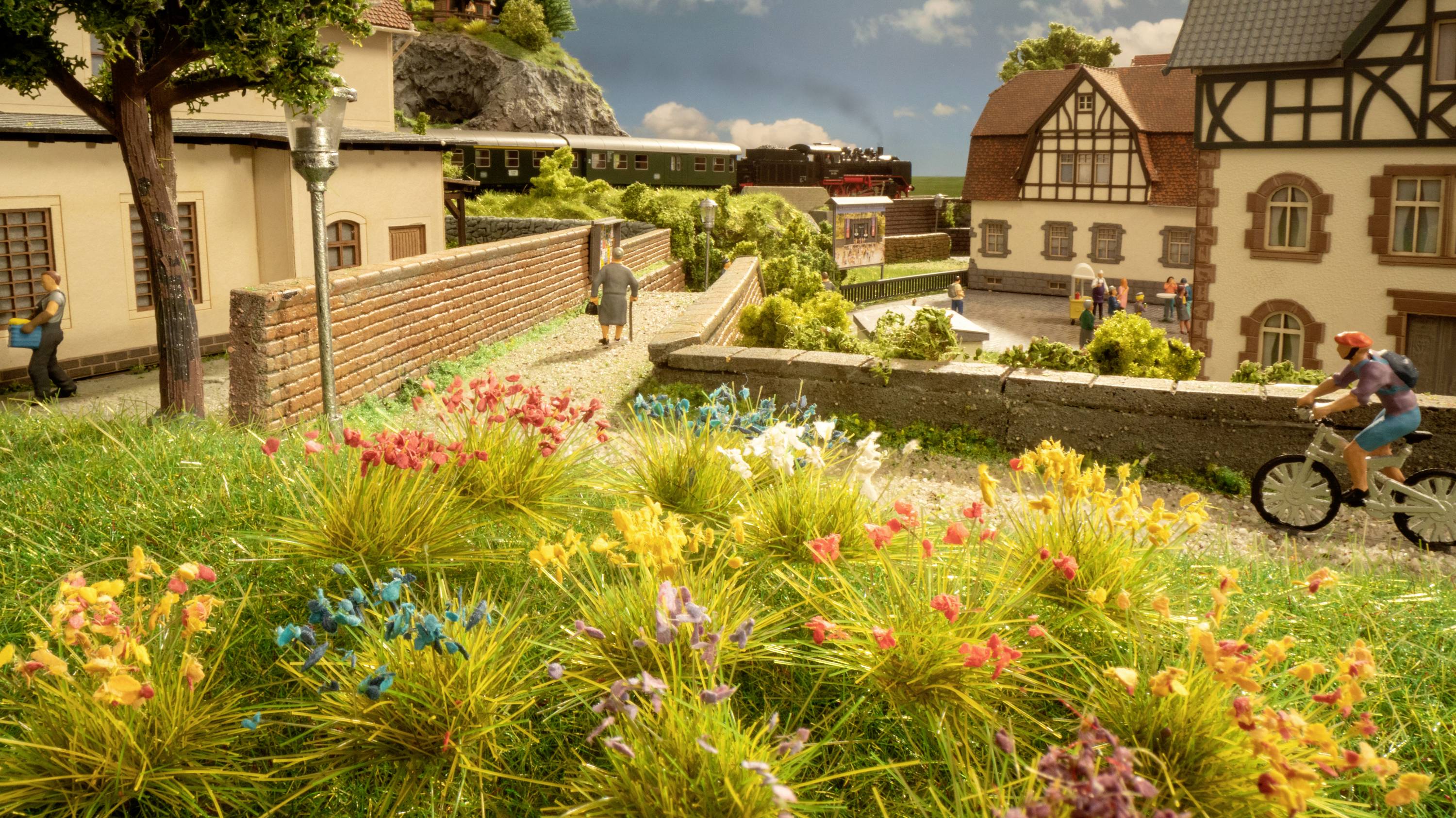 Eine Modelleisenbahnlandschaft mit bunten Blumen im Vordergrund, einem vorbeifahrenden Zug im Hintergrund und Figuren bei Häusern.