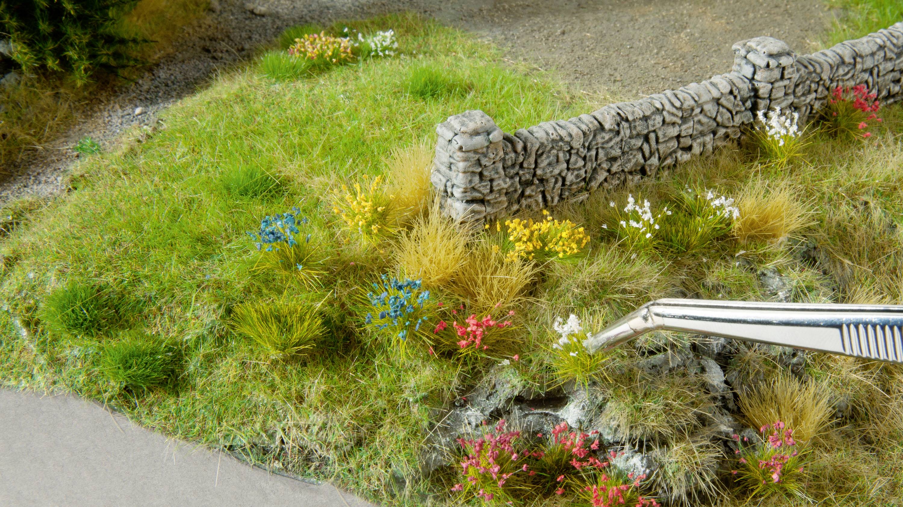 Eine Modelllandschaft zeigt eine kleine Wiese mit bunten Blumen und einer niedrigen Steinmauer. Eine Pinzette platziert Pflanzen.