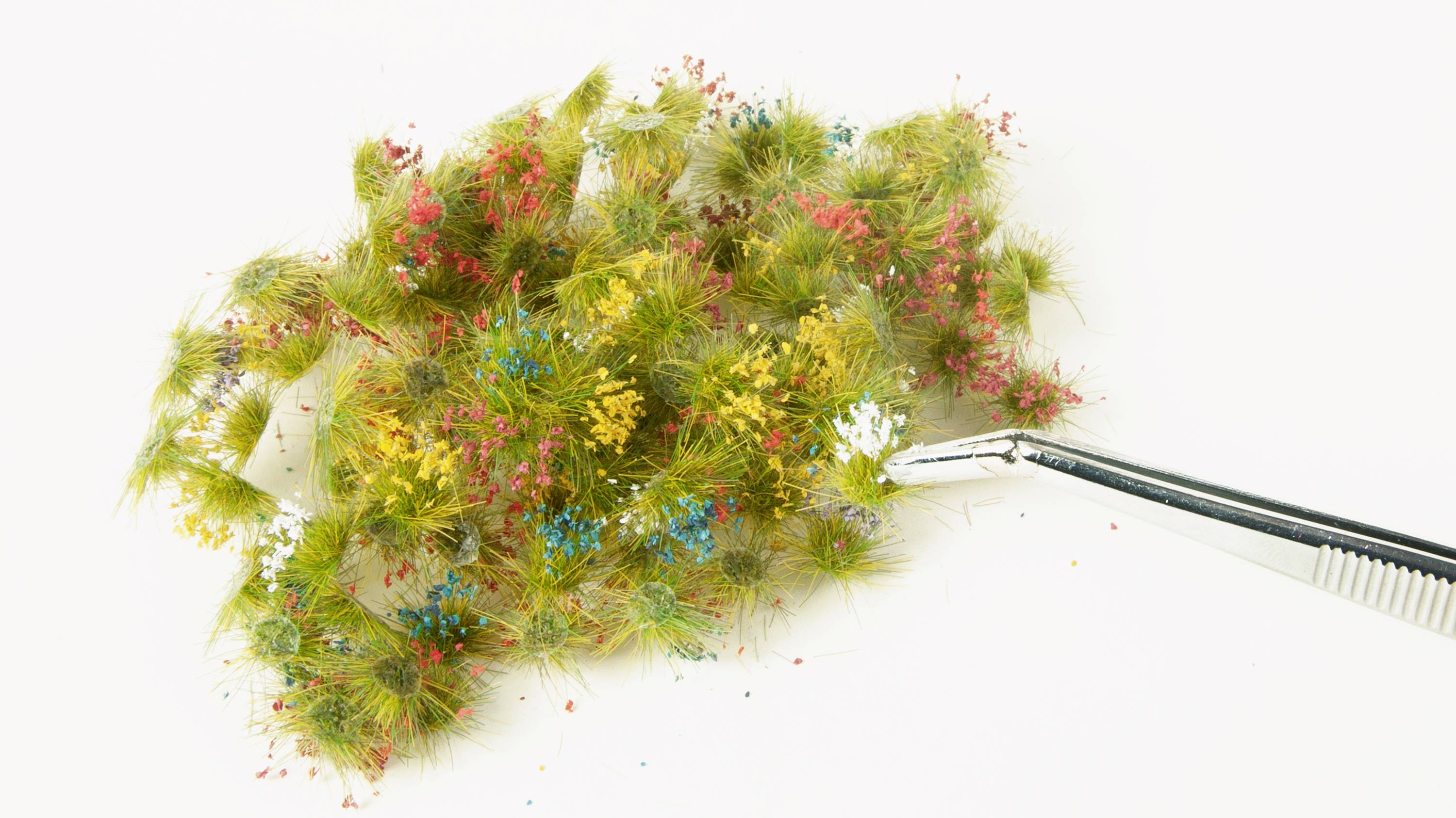 Eine Pinzette hält ein kleines Bündel aus grünem, gelbem, rotem und blauem Bastelmoos auf einem weißen Hintergrund.