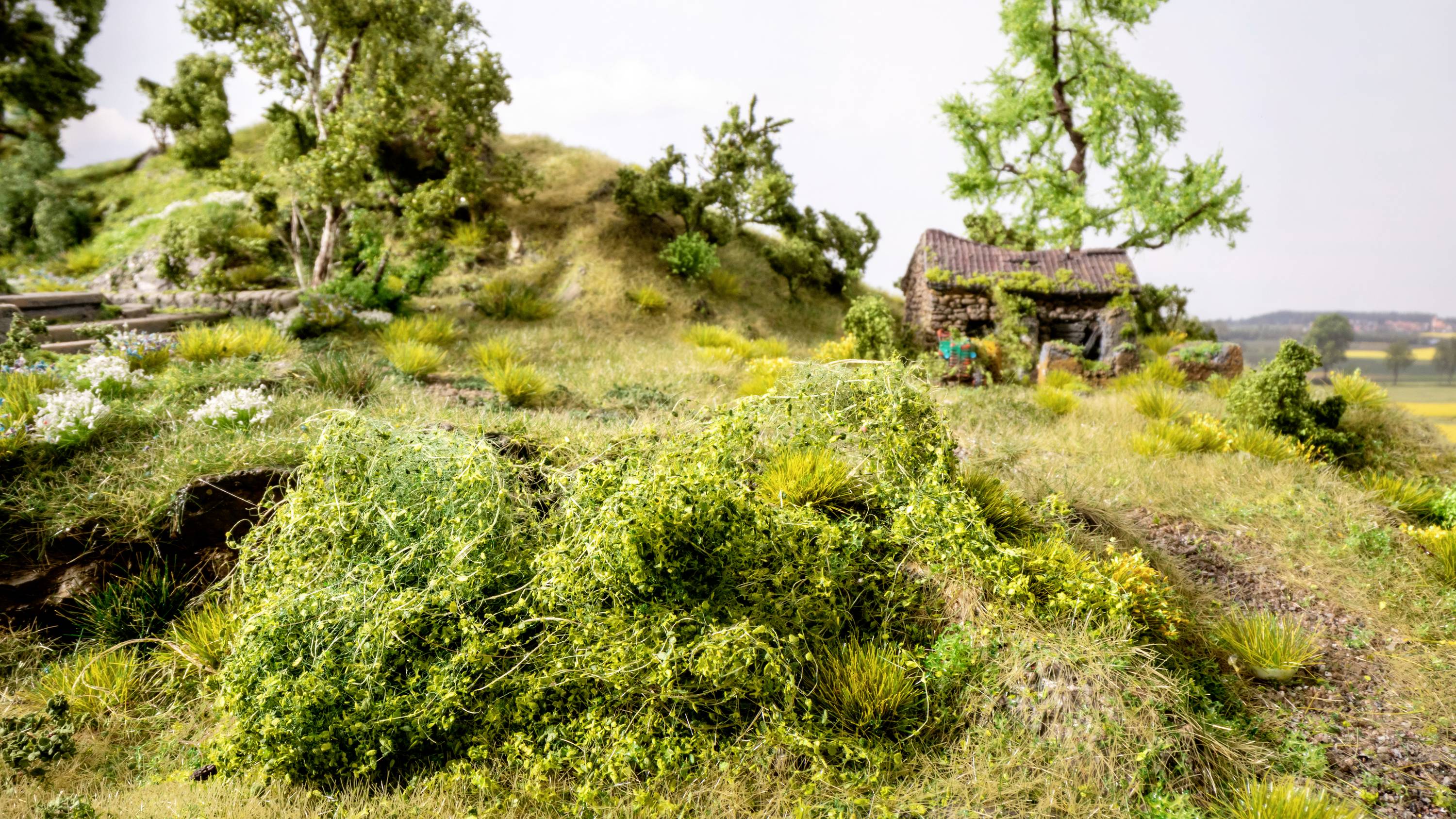 Eine Miniaturlandschaft mit einem kleinen Steinhaus, grünen Büschen, Bäumen und einem kiesigen Weg. Der Himmel ist klar.