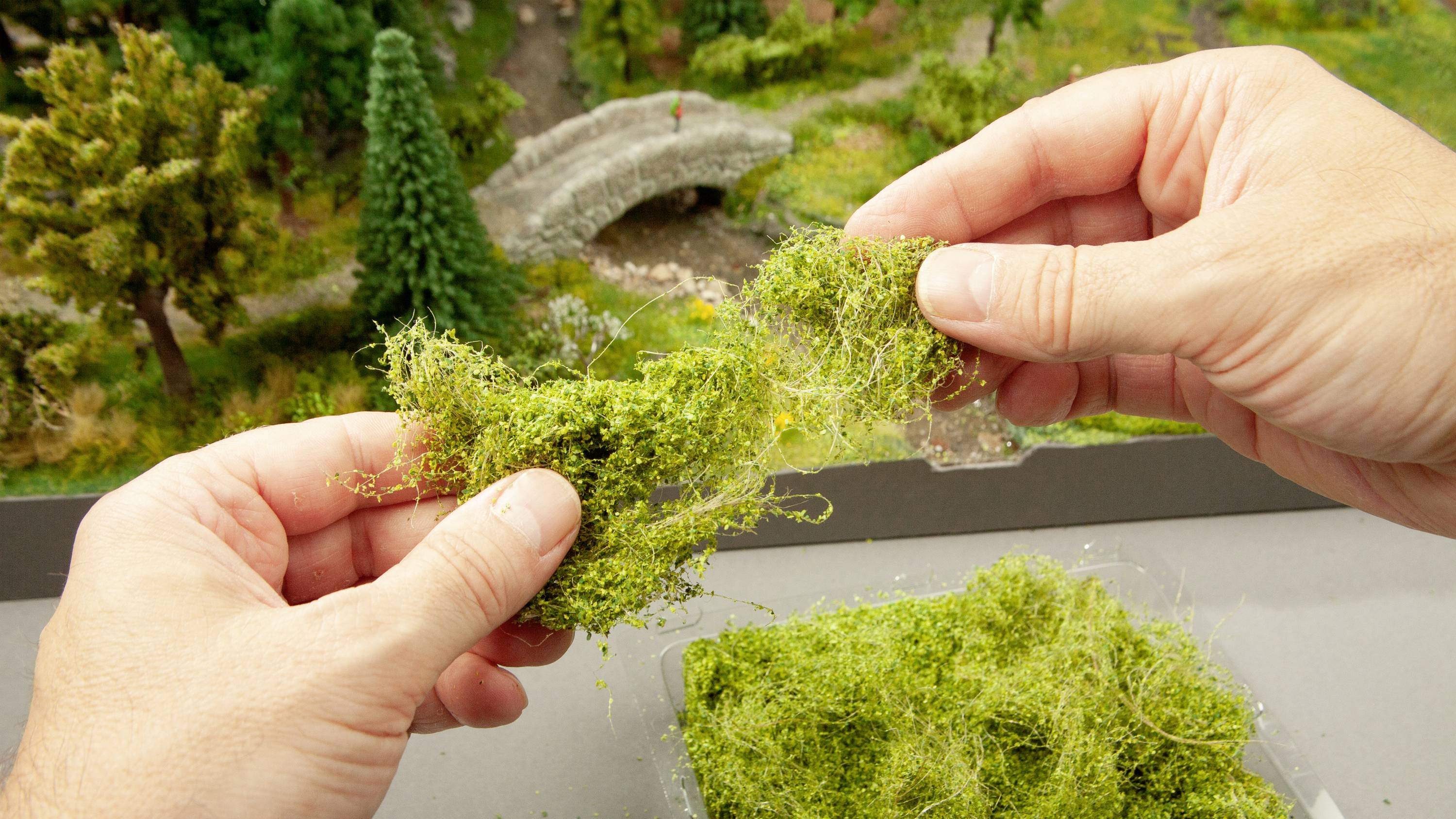 Zwei Hände formen grüne Modellbau-Landschaftsbegrünung vor einem Miniaturlandschaftsmodell mit einem kleinen Steinbogen.