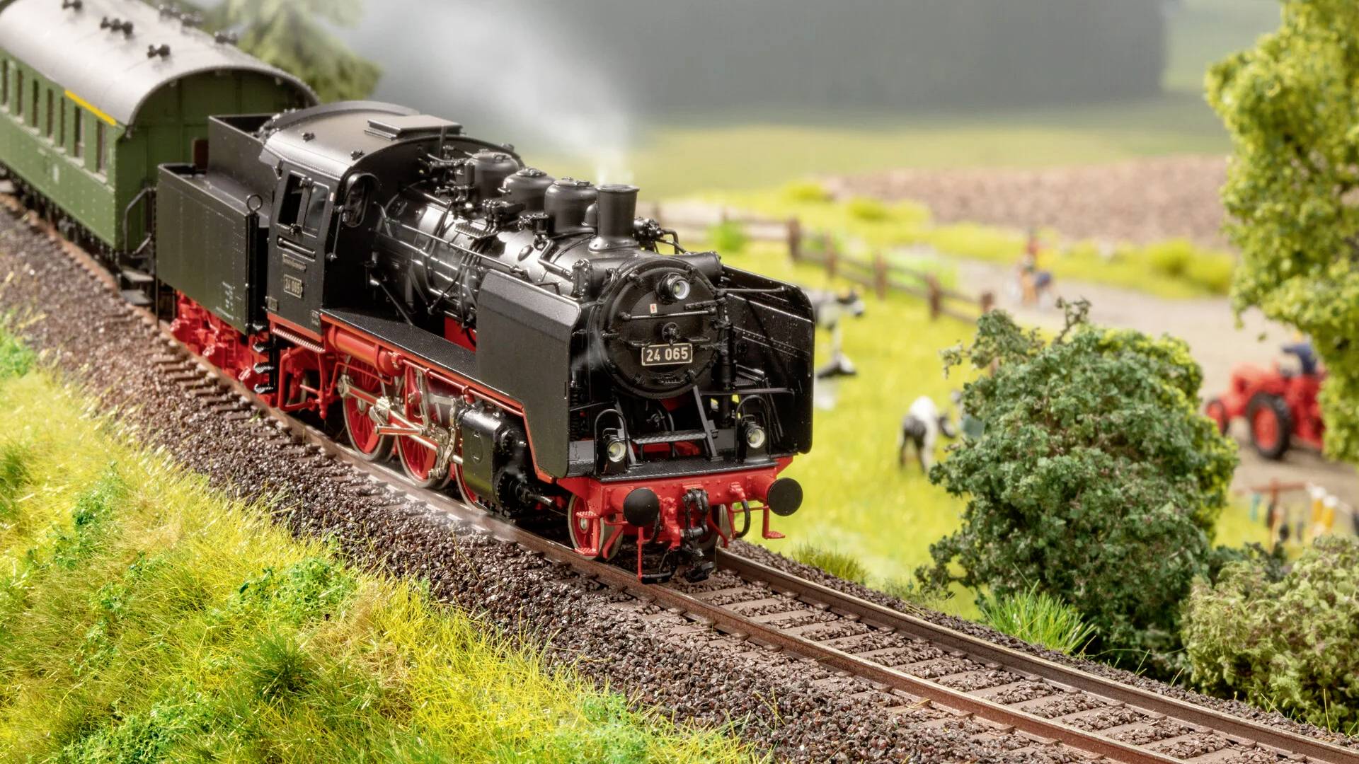 Modelleisenbahn-Szene mit einer Dampflokomotive, die auf einer Gleisstrecke fährt, umgeben von grüner Landschaft und Bäumen.