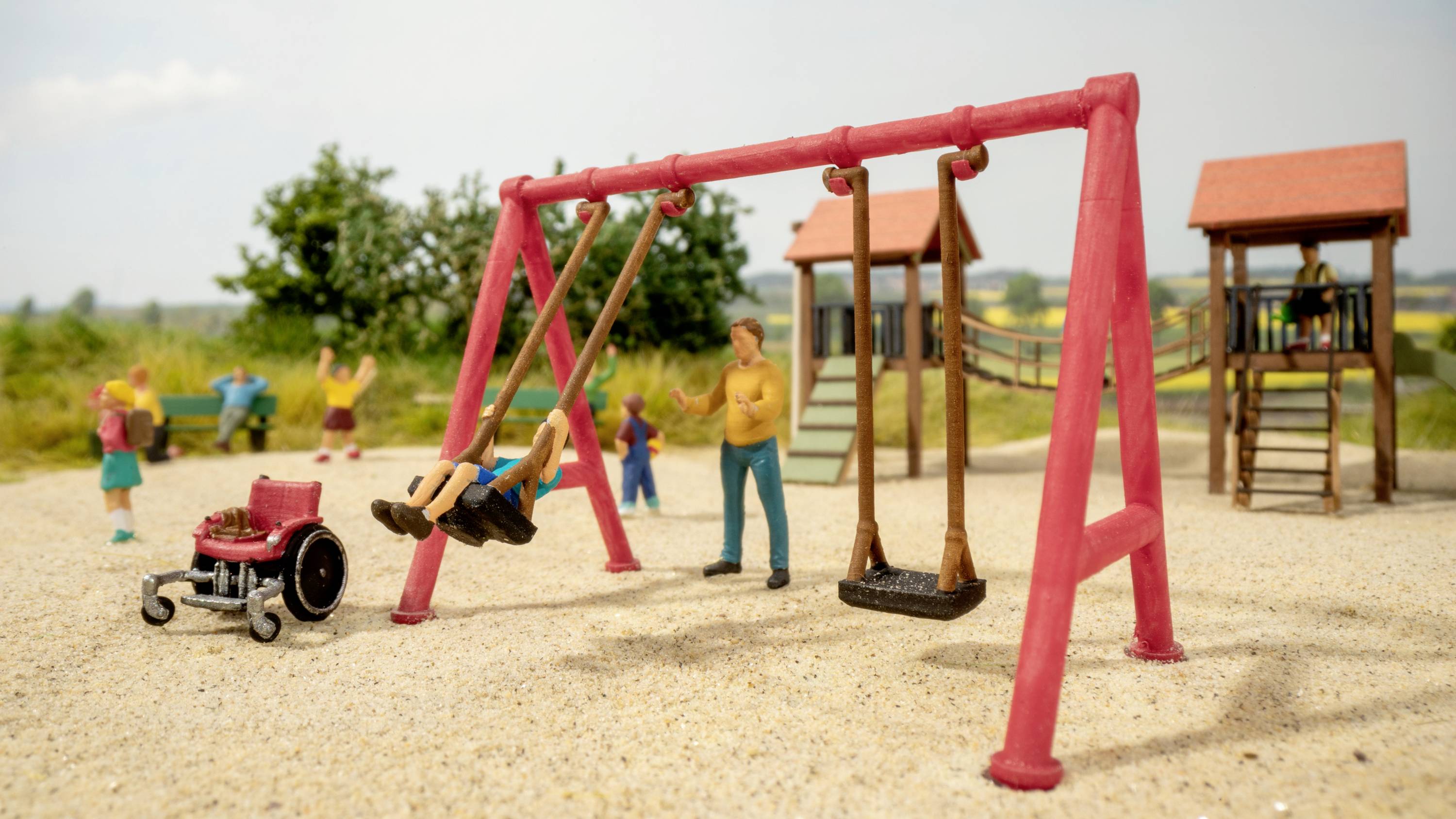 Spielplatzszene mit Kindern, die auf einer Schaukel spielen, ein rollstuhlgerechter Zugang ist sichtbar. Im Hintergrund sind ein Klettergerüst und Bäume.