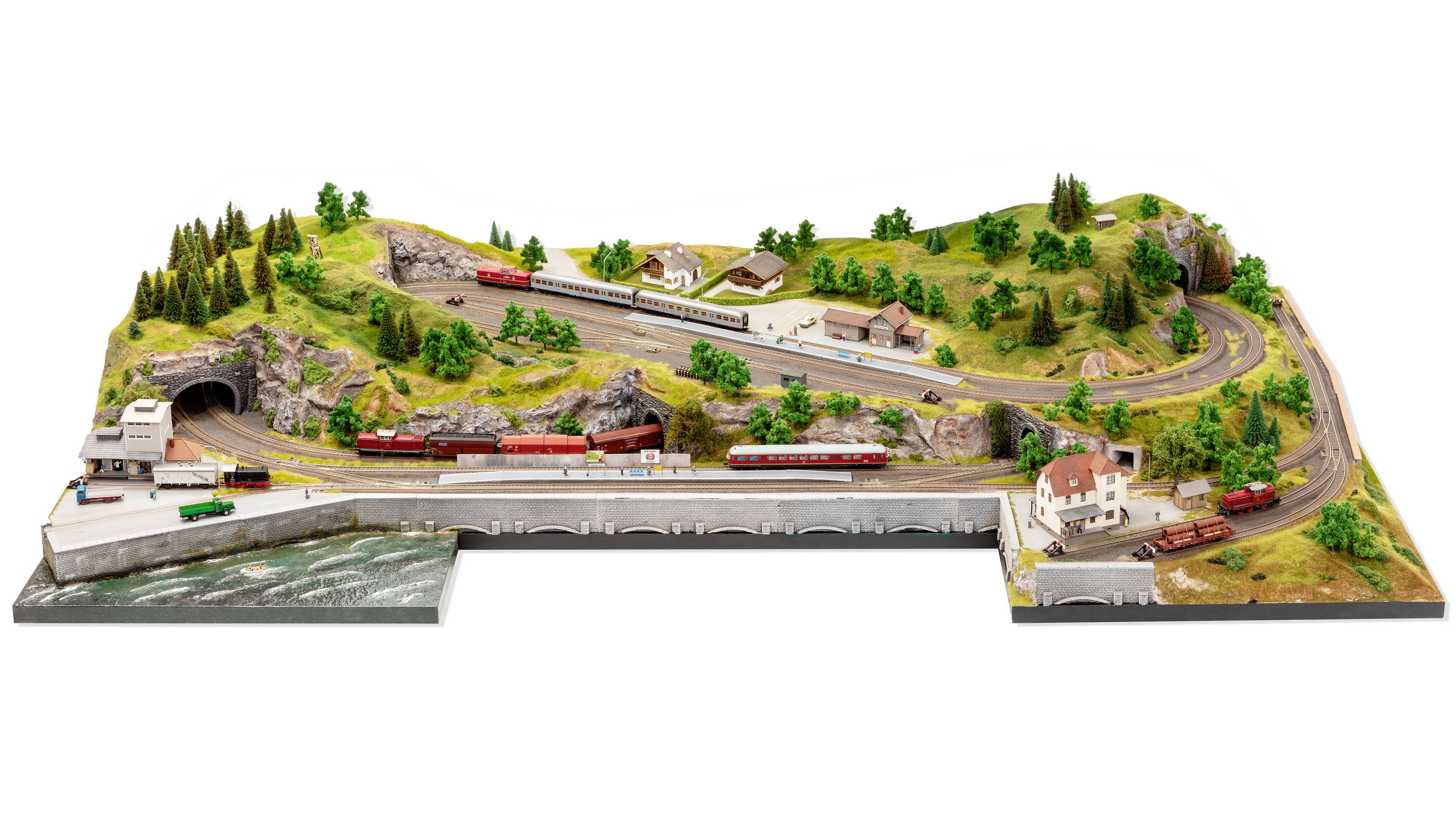 Eine Modelleisenbahnlandschaft mit einem Zug, der über Brücken und durch einen Tunnel fährt. Umgeben von grünen Hügeln und Gebäuden.