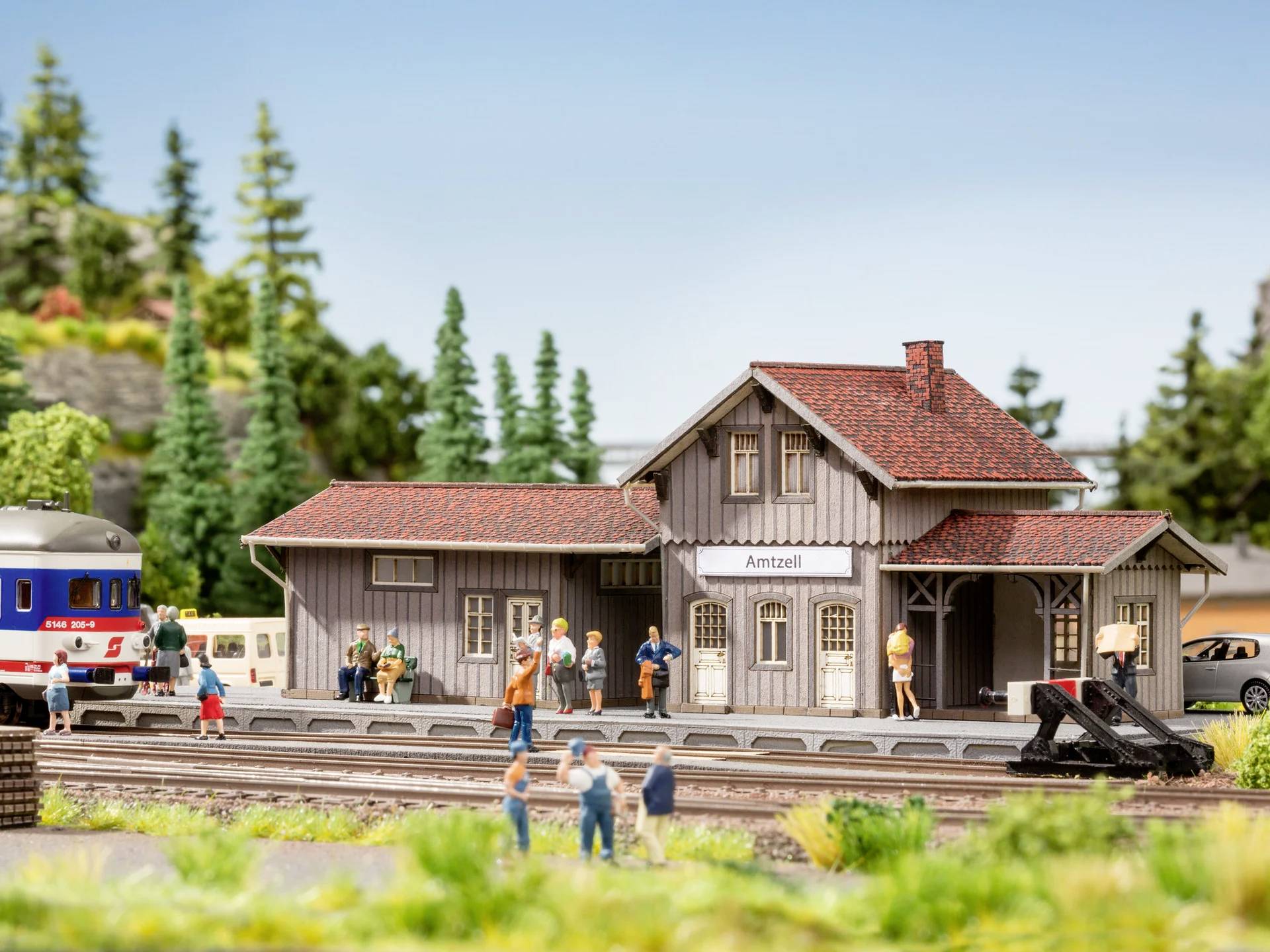 Miniatur-Bahnhofsszene mit Menschen auf dem Bahnsteig; Gebäude mit Schild 'Amtzell', umgeben von Modellbäumen und Gleisen.
