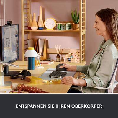 Eine Frau sitzt an einem Schreibtisch, entspannt den Oberkörper und betrachtet den Bildschirm. Im Hintergrund sind Regale mit Gegenständen.