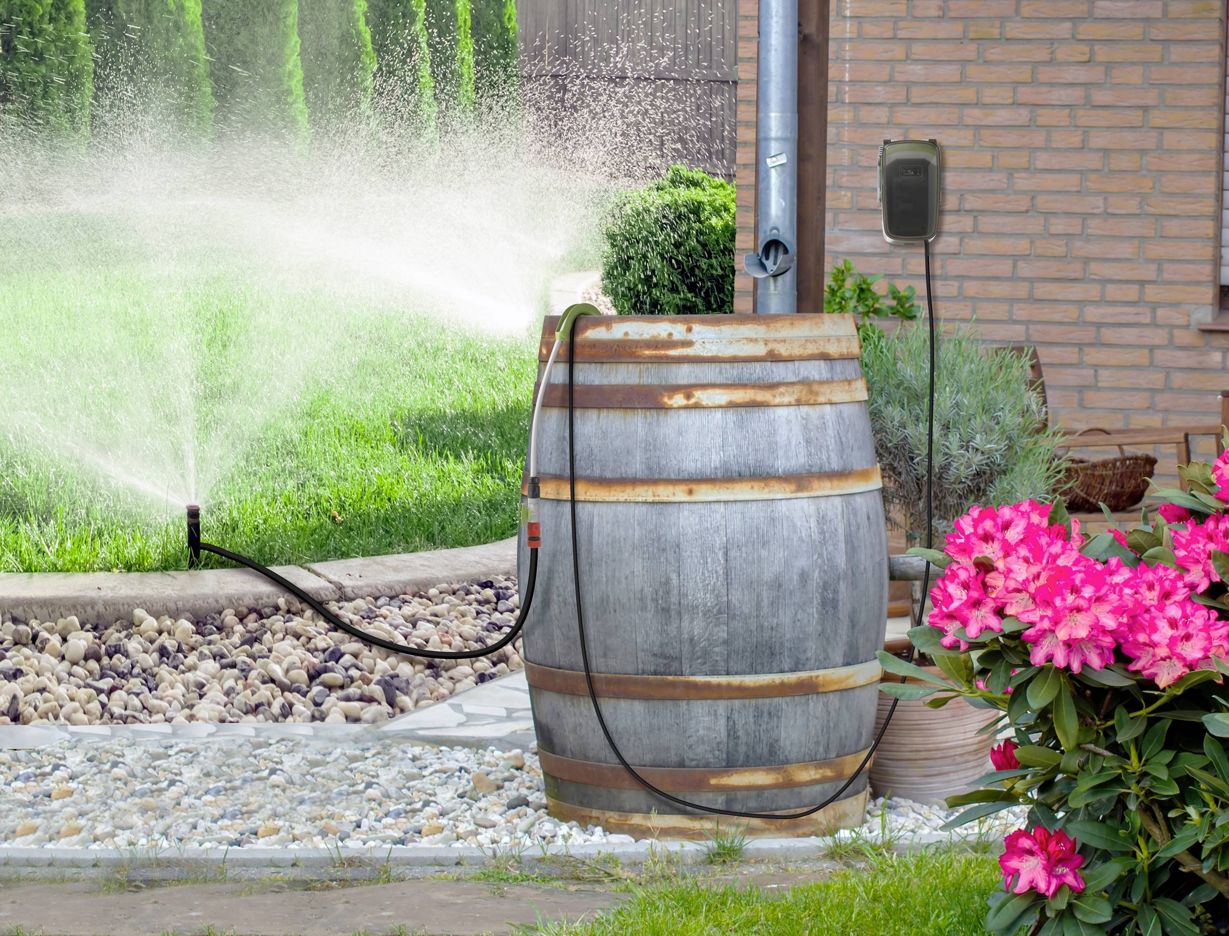 Ein Fass sammelt Regenwasser, das zur Bewässerung eines Gartens mit Rasen und Blumen verwendet wird. Ein Sprinkler verteilt das Wasser.