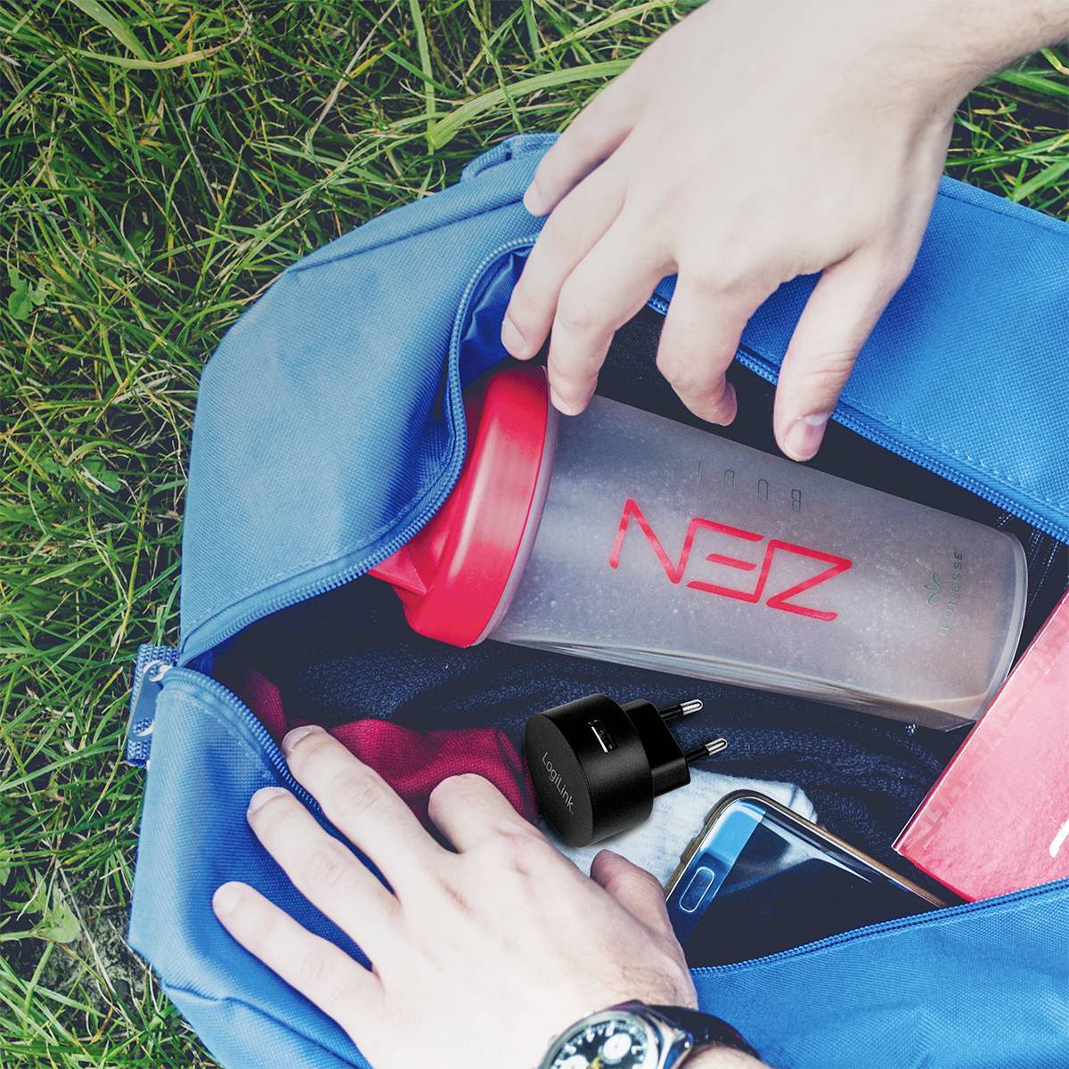 Person packt einen Sportbecher und ein Ladegerät in eine blaue Tasche. Ein rotes Buch und ein Smartphone sind ebenfalls sichtbar.