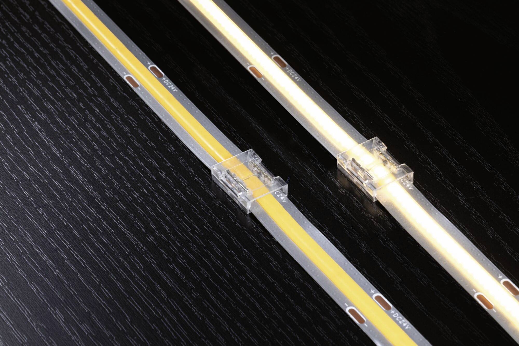 Zwei LED-Streifen auf einer dunklen Oberfläche, verbunden mit transparenten Clips. Einer leuchtet, der andere ist ausgeschaltet.