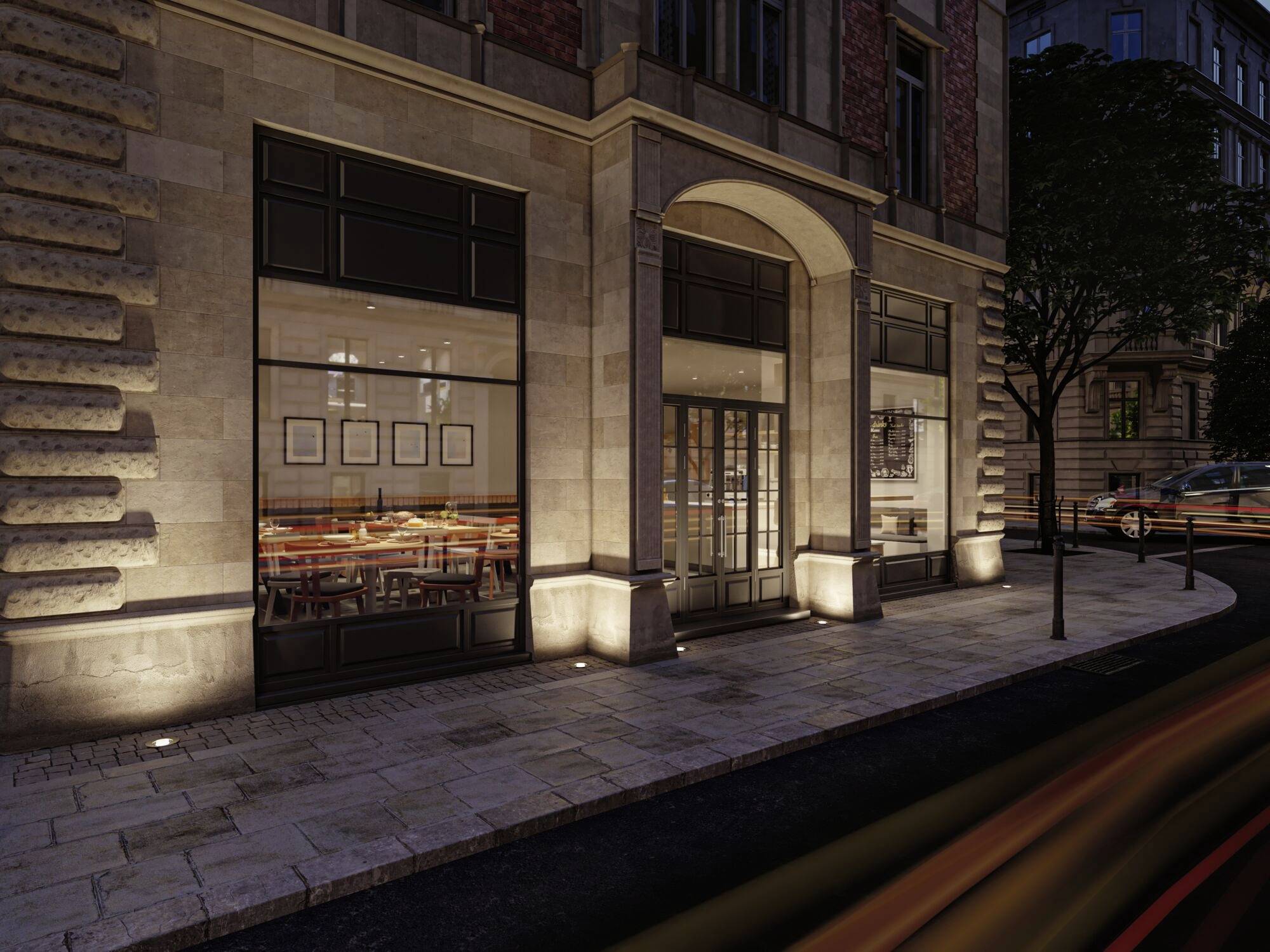 Außenansicht eines Restaurants bei Nacht mit beleuchteten Fenstern und einem Baum rechts, Umgebung eher leer und ruhig.