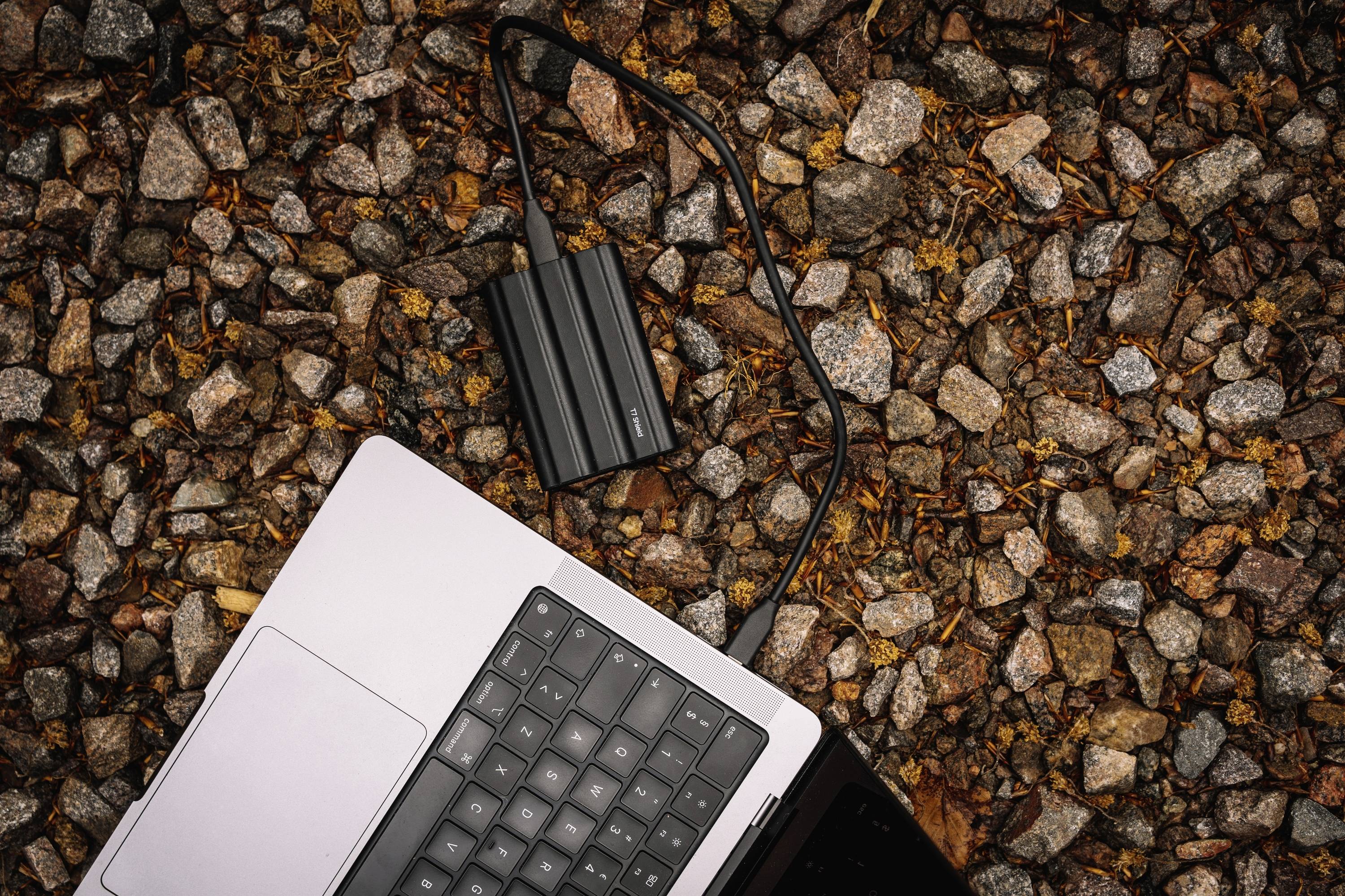 Ein Laptop liegt auf einem Kiesboden. Ein schwarzes Kabel ist angeschlossen und führt zu einem externen Gerät neben dem Laptop.
