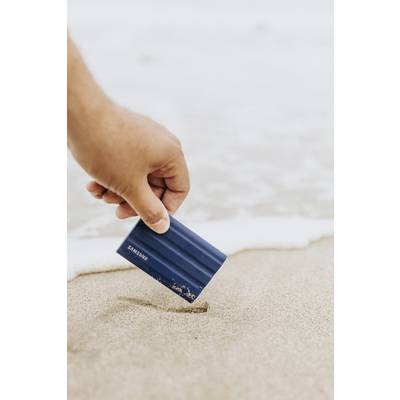 Eine Hand hebt ein kleines blaues Gerät aus dem Sand an einem Strand. Im Hintergrund ist die sanfte Brandung zu sehen.
