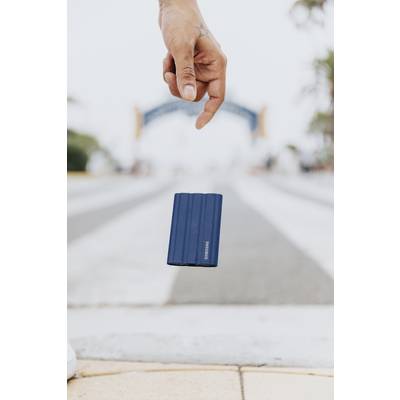 Eine Hand hält eine kleine, blaue, rechteckige Festplatte über einer unscharfen, breiten Straße mit Bäumen und einem Tor im Hintergrund.