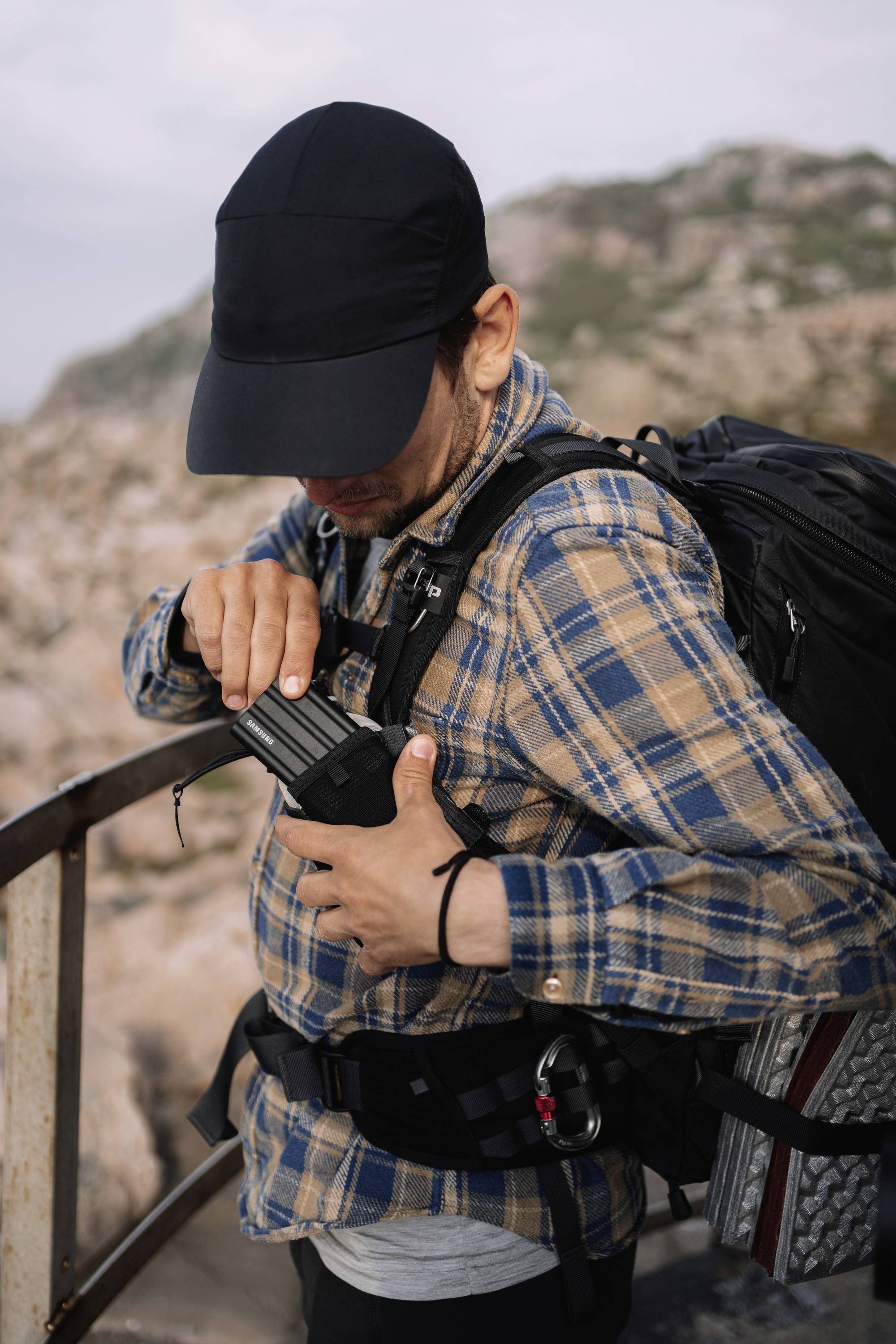 Ein Mann mit Basecap und Rucksack zieht ein Objekt aus seiner Tasche. Er steht auf einer Aussichtsplattform mit felsiger Landschaft im Hintergrund.