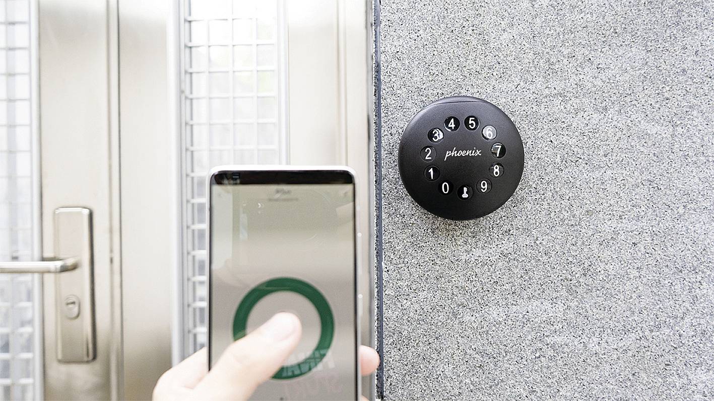 Eine Hand hält ein Smartphone vor einem digitalen Türschloss, das Zahlen zeigt.