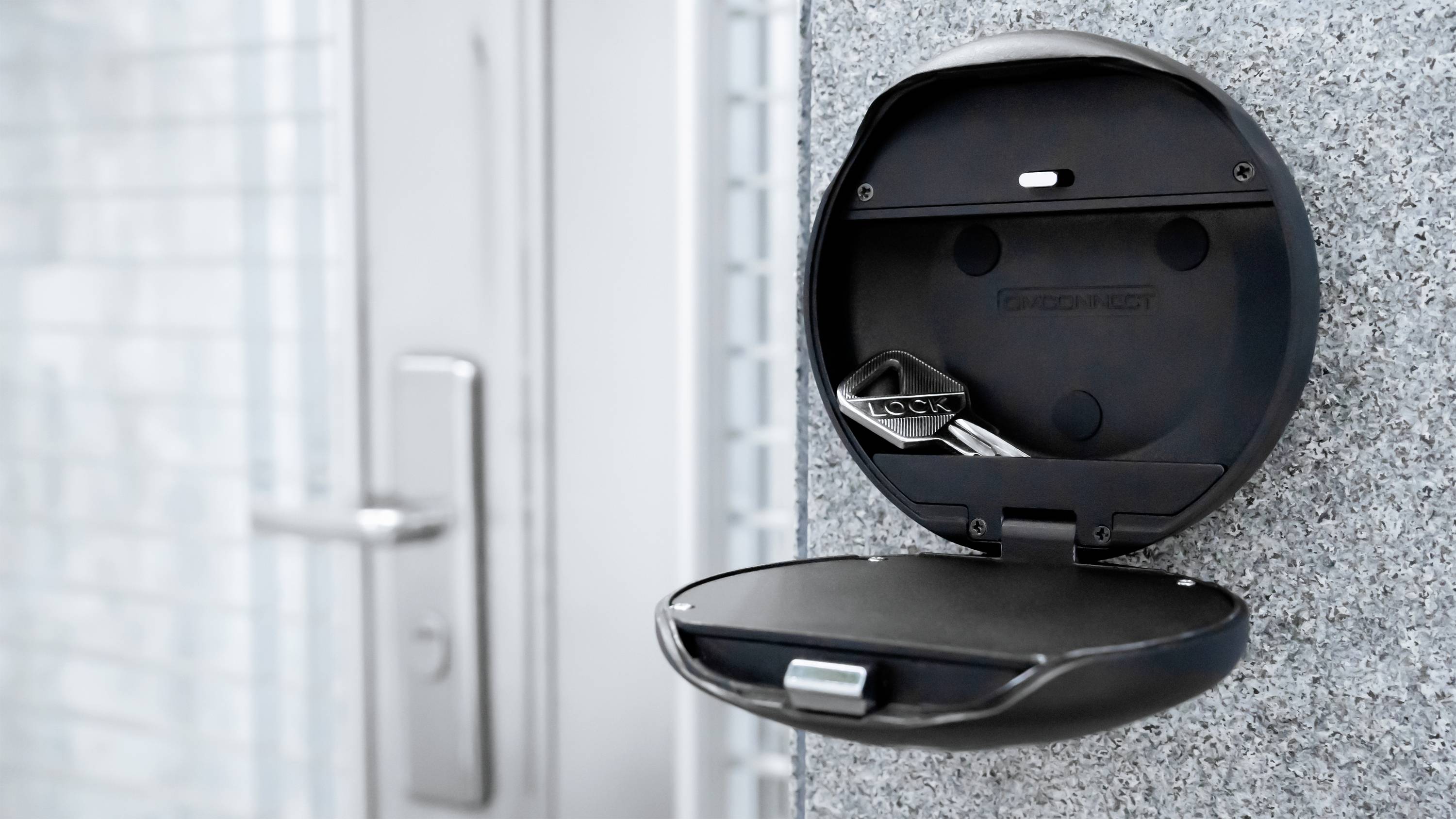 Schlüsselbox an einer Wand montiert, geöffnet und leer bis auf einen einzelnen Metallschlüssel, neben einer verschlossenen Tür.