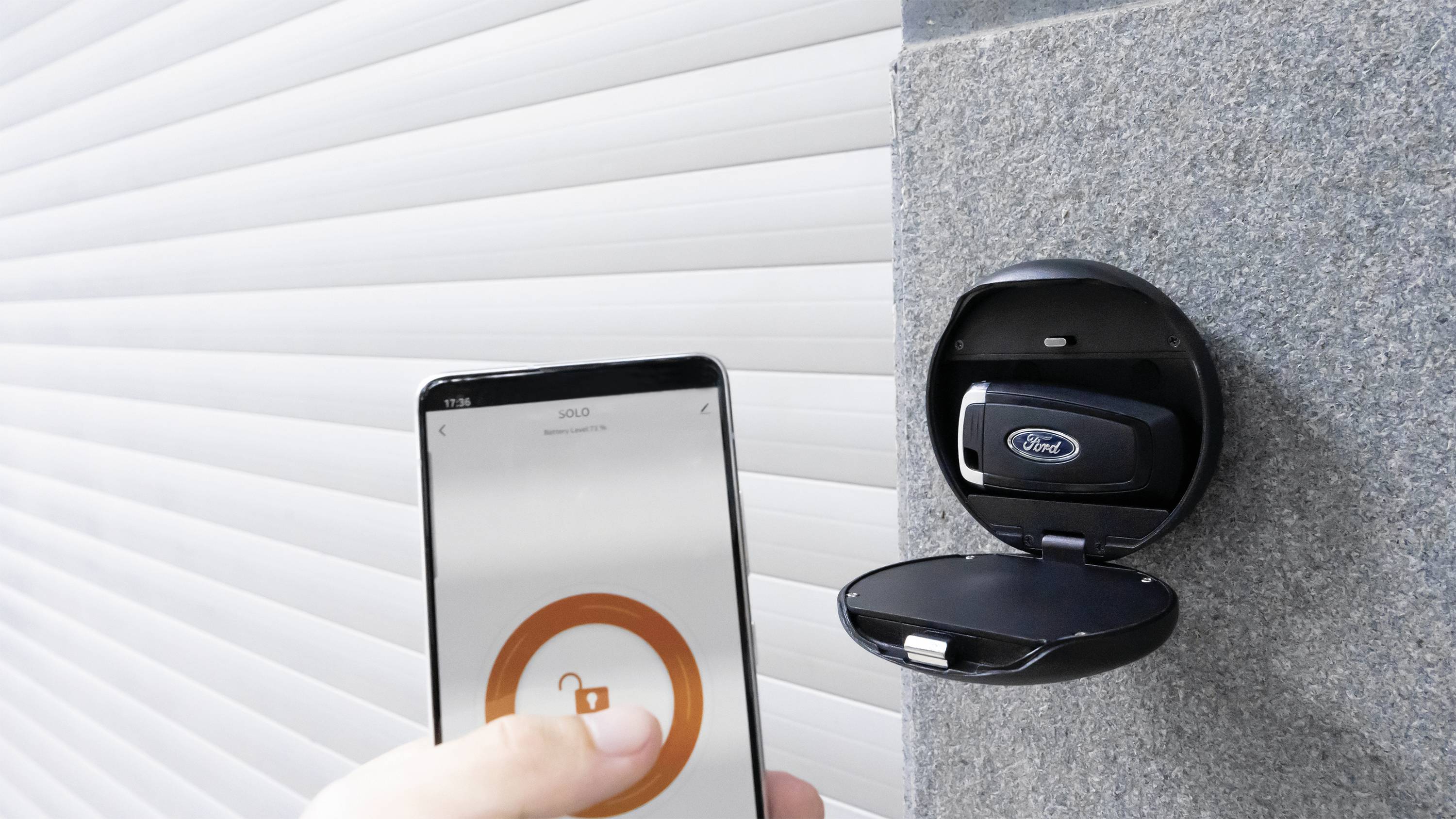 Eine Hand hält ein Smartphone vor einem geöffneten Schlüsselsafe, der einen Ford-Autoschlüssel enthält, vor einem Rolltor.