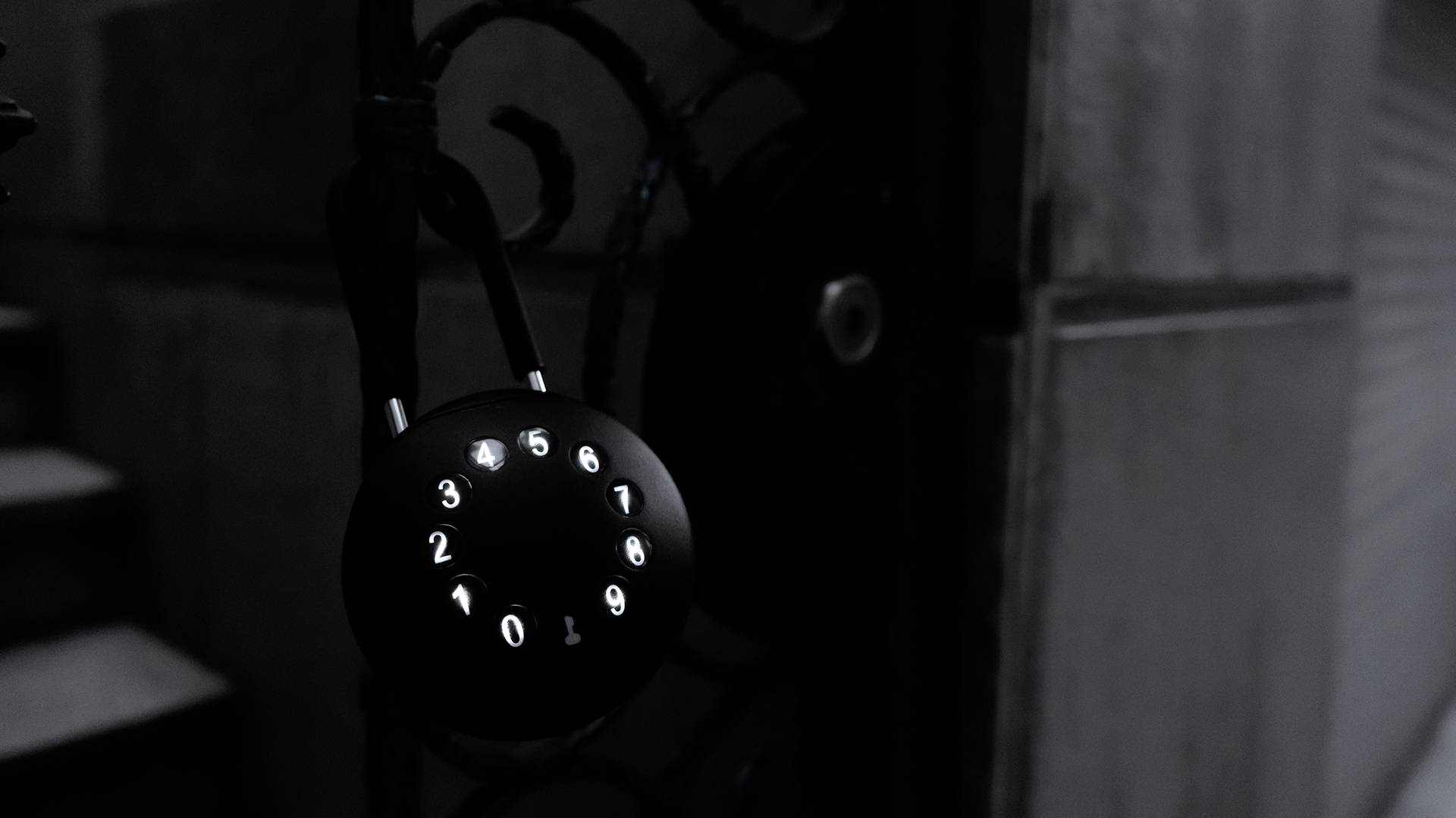 Ein schwarzes Zahlenschloss mit leuchtenden Ziffern von 0 bis 9 hängt an einem metallenen Tor. Es ist Nacht, Umgebungslicht ist schwach.