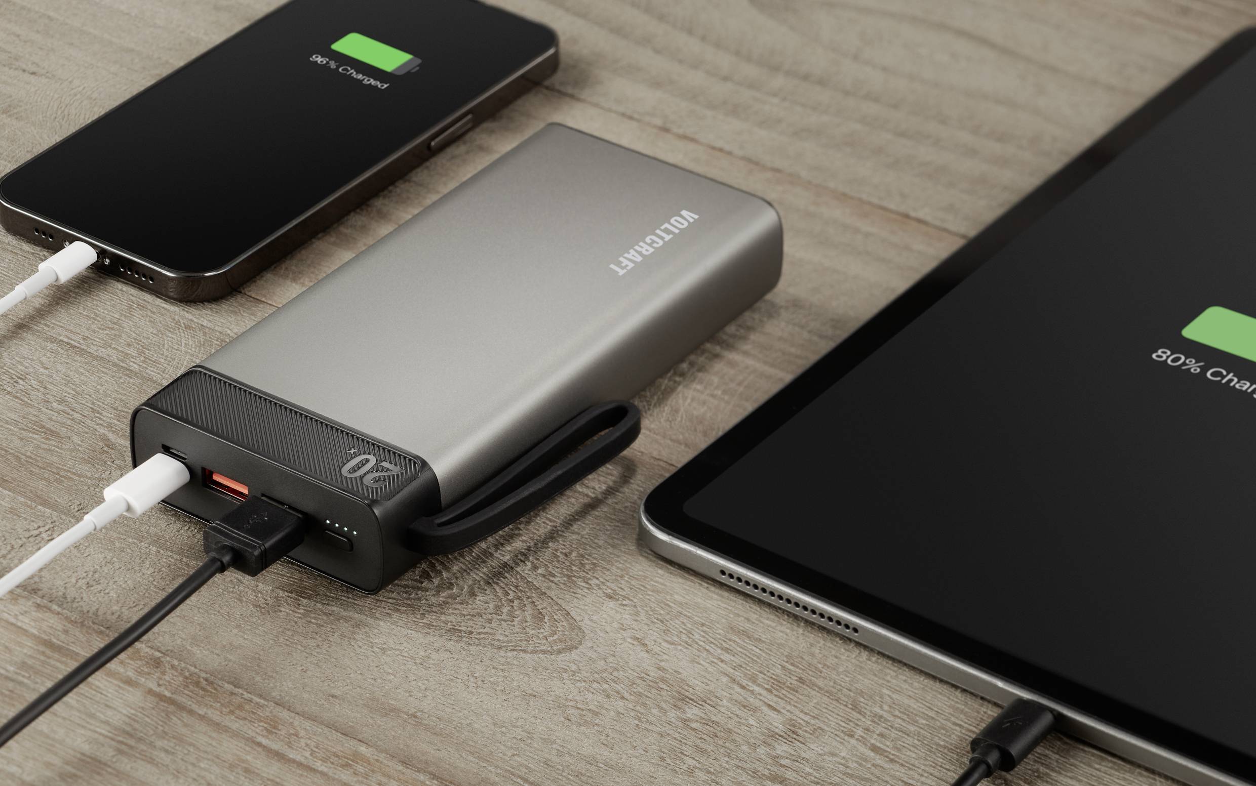 Ein Smartphone, eine Powerbank und ein Tablet liegen nebeneinander auf einem Tisch. Das Smartphone und das Tablet werden aufgeladen.
