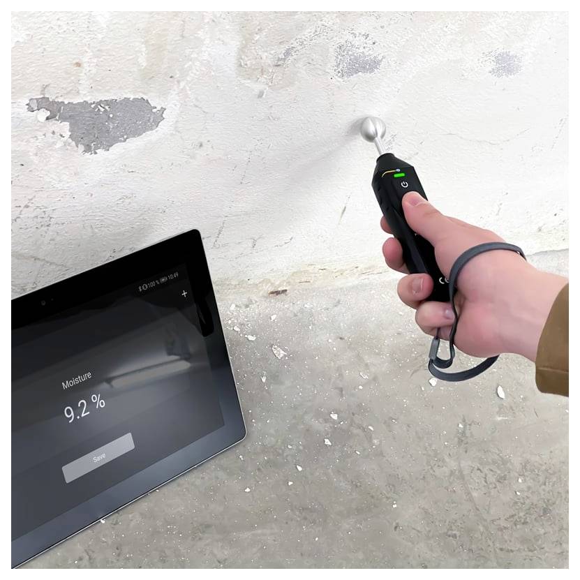 Tragbares Feuchtemessgerät, das eine feuchte Betonwand misst, während auf einem Tablet „Feuchtigkeit 9,2 %" angezeigt wird.