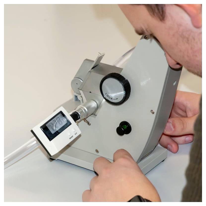 Person passt ein optisches Tischinstrument mit digitaler Anzeige und Fokussierknopf an, während sie/er eine Linsen- oder Röhreneinheit untersucht.