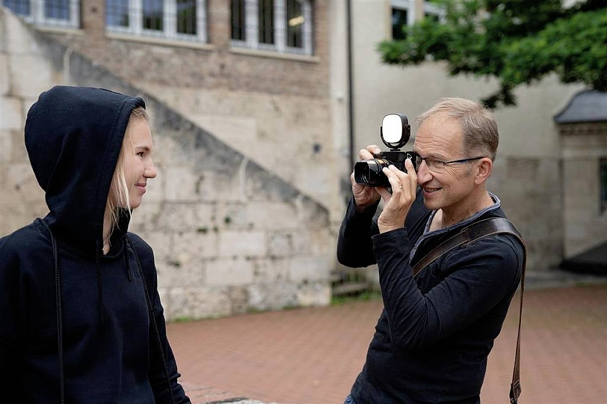 Ein Mann fotografiert eine Person, die einen schwarzen Hoodie trägt. Im Hintergrund sind Gebäude und Bäume zu sehen.