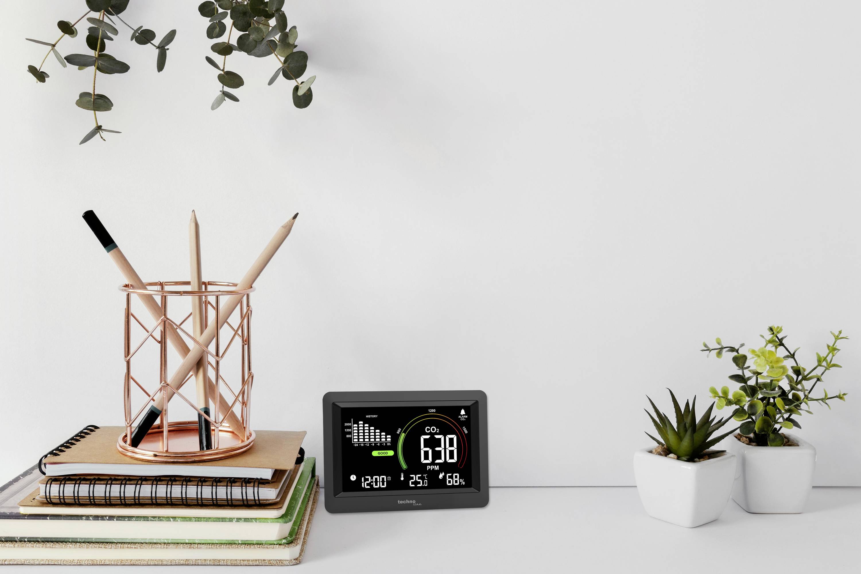 Ein Tisch mit Notizbüchern und einem Stifthalter links. Rechts ein kleines digitales Display zeigt CO2-Gehalt, Temperatur und Luftfeuchtigkeit an. Daneben stehen zwei kleine Pflanzen.