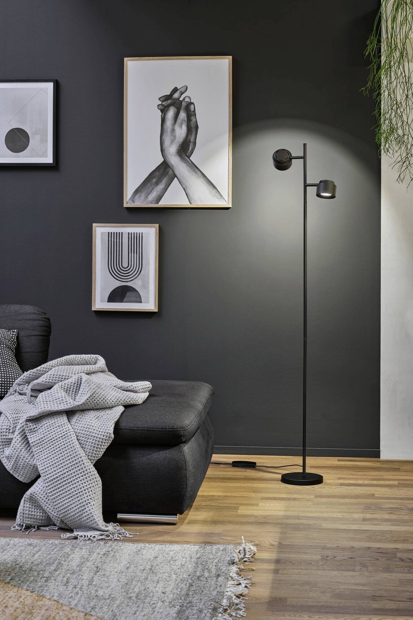 Ein modernes Wohnzimmer mit einer Stehlampe, grauem Sofa und drei Kunstwerken an der Wand. Holzfußboden und graue Akzente schaffen eine gemütliche Atmosphäre.