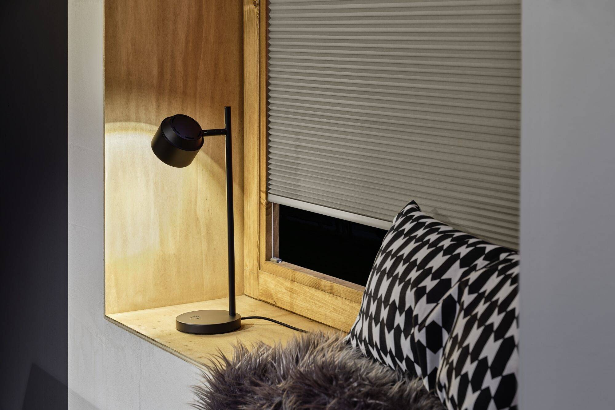 Eine Tischlampe beleuchtet eine Fensterbank mit Kissen und einem Pelzbezug. Das Fenster ist mit einem Rollladen teilweise abgedunkelt.